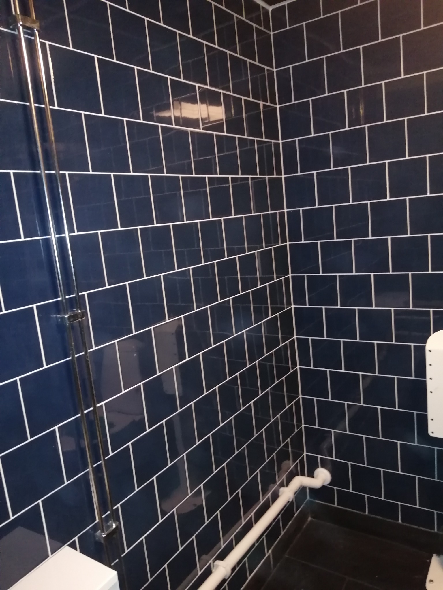 Narożnik kabiny prysznicowej wyłożony ciemnymi, prostokątnymi płytkami z białą fugą, widoczna chromowana rura prysznicowa i biała rura kanalizacyjna przy podłodze.