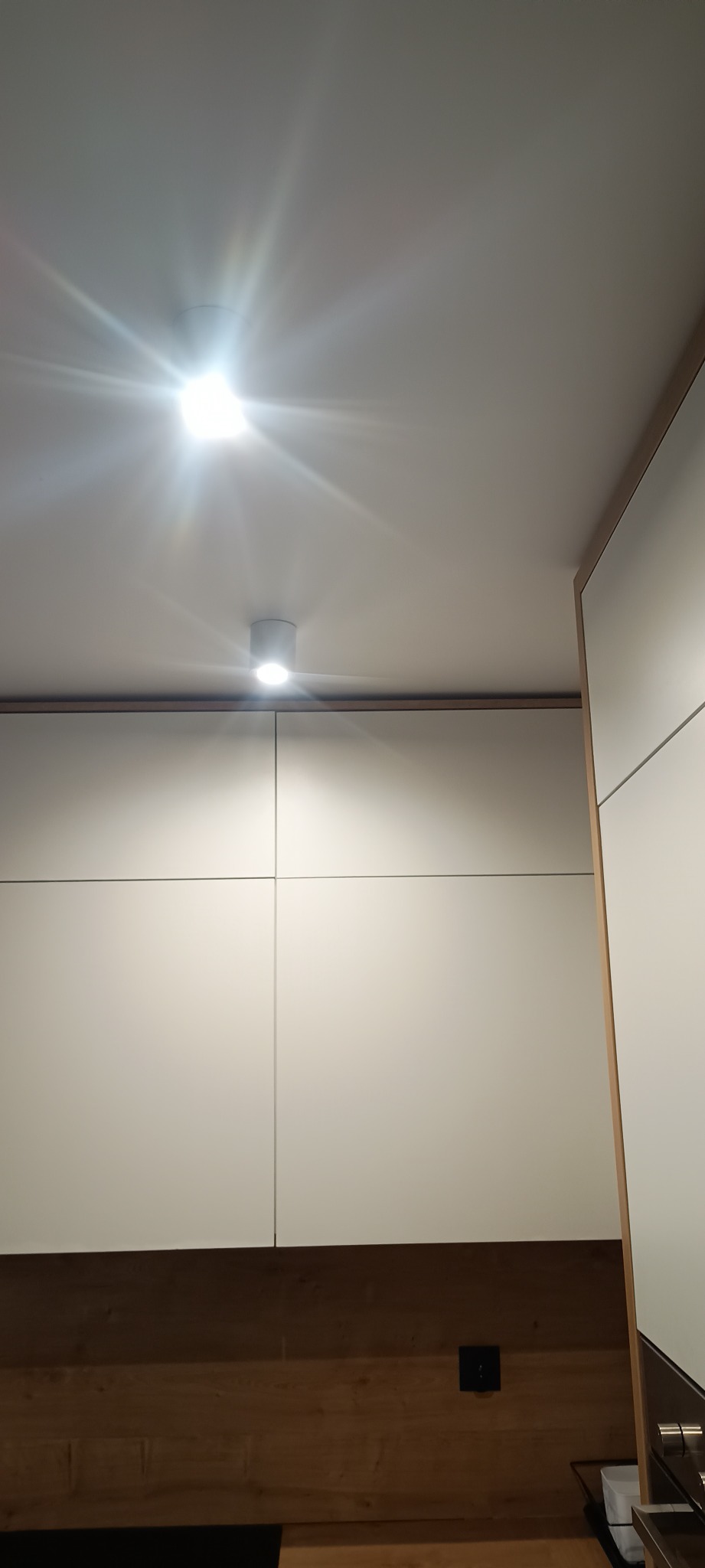 Dwa punktowe reflektory sufitowe LED włączone, oświetlające białą zabudowę meblową z drewnianym akcentem, widok od dołu.