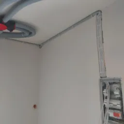 Szare rury karbowane prowadzone po suficie i ścianie, widoczna skrzynka z bezpiecznikami z podłączonymi przewodami, ściany w stanie surowym.