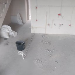 Tjg - Surowy stan wykończeniowy wnętrza: widoczne przewody elektryczne w ścianach, przygotowane punkty pod gniazdka, resztki gruzu na podłodze, wiadro i worki z materiałami budowlanymi obok schodów.