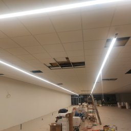 Tjg - Wnętrze komercyjne w trakcie montażu oświetlenia liniowego LED w suficie podwieszanym, widoczne braki w panelach sufitowych i rozłożone materiały instalacyjne na podłodze.