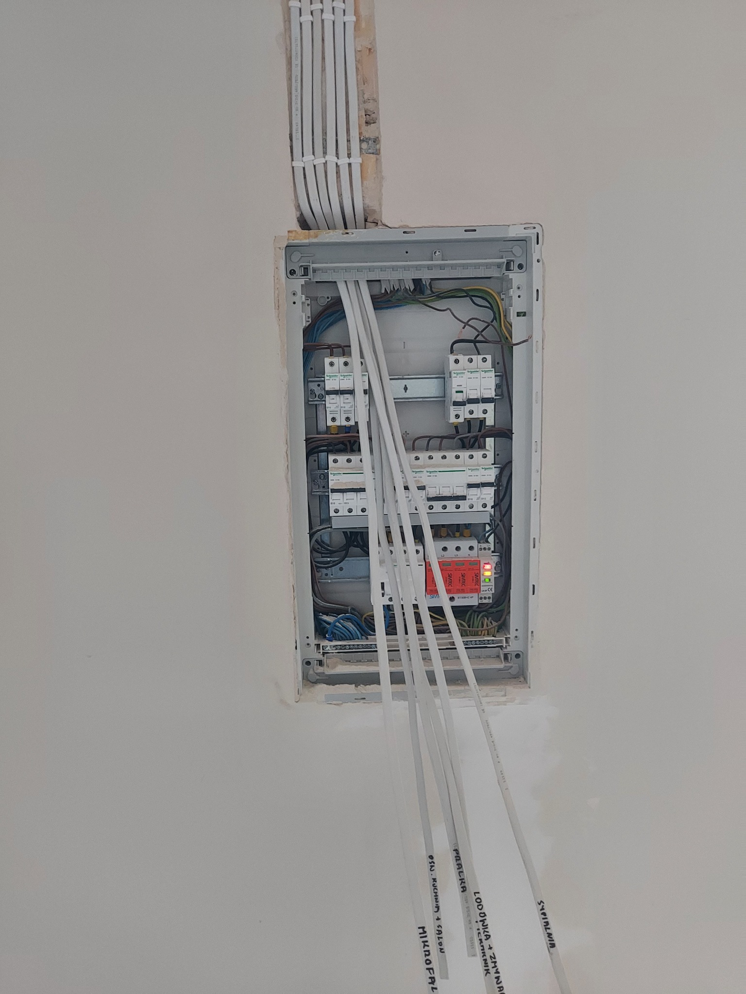 Rozdzielnia elektryczna w trakcie montażu, widoczne przewody z opisami (sypialnia, lodówka, pralka, mikrofalówka) wychodzące ze ściany, zabezpieczenia różnicowoprądowe i nadprądowe wewnątrz, kable...