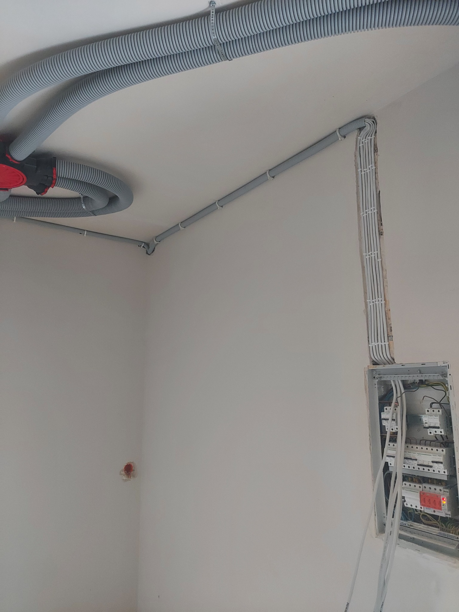 Szare rury karbowane prowadzone po suficie i ścianie, widoczna skrzynka z bezpiecznikami z podłączonymi przewodami, ściany w stanie surowym.