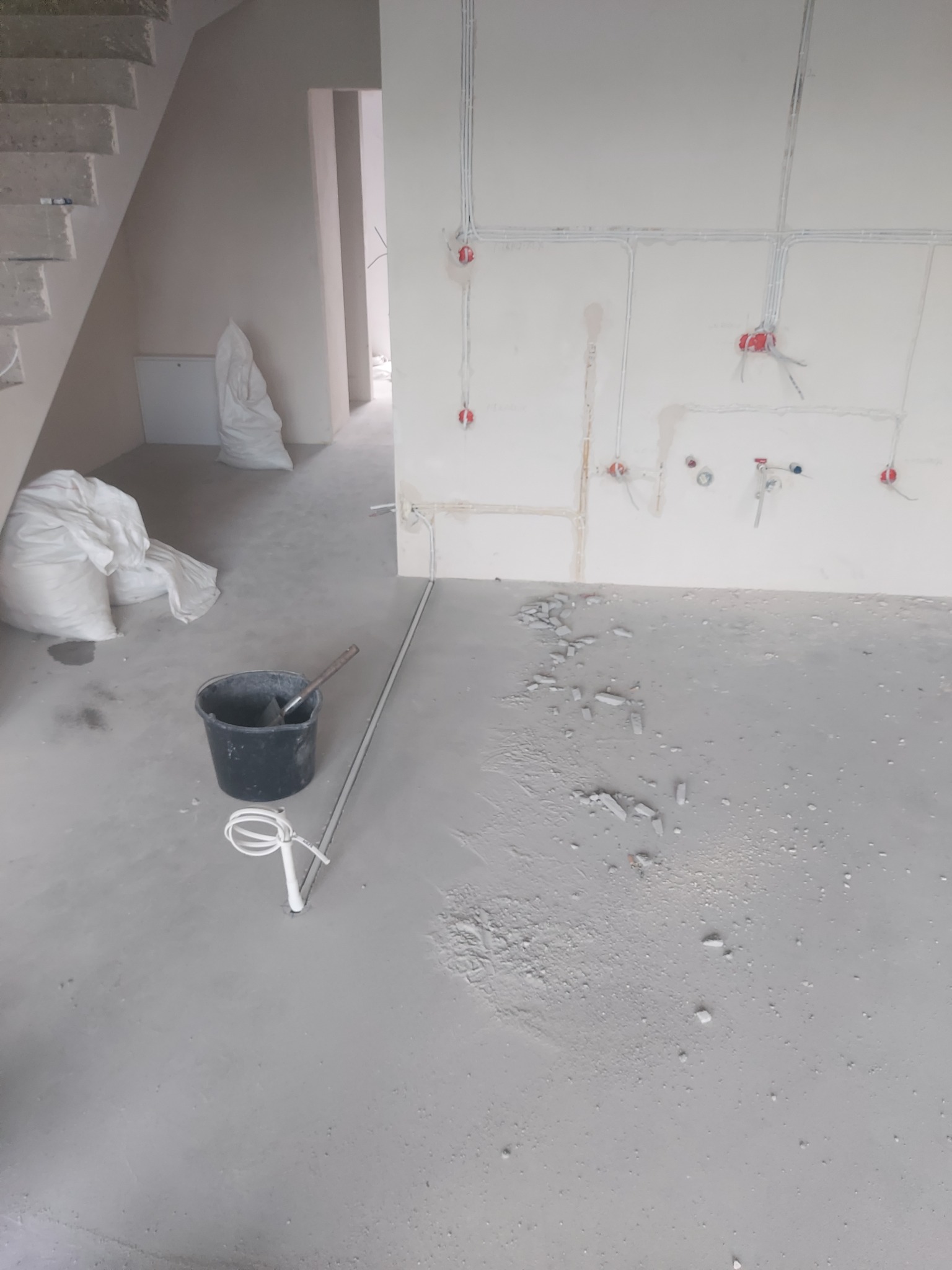 Surowy stan wykończeniowy wnętrza: widoczne przewody elektryczne w ścianach, przygotowane punkty pod gniazdka, resztki gruzu na podłodze, wiadro i worki z materiałami budowlanymi obok schodów.