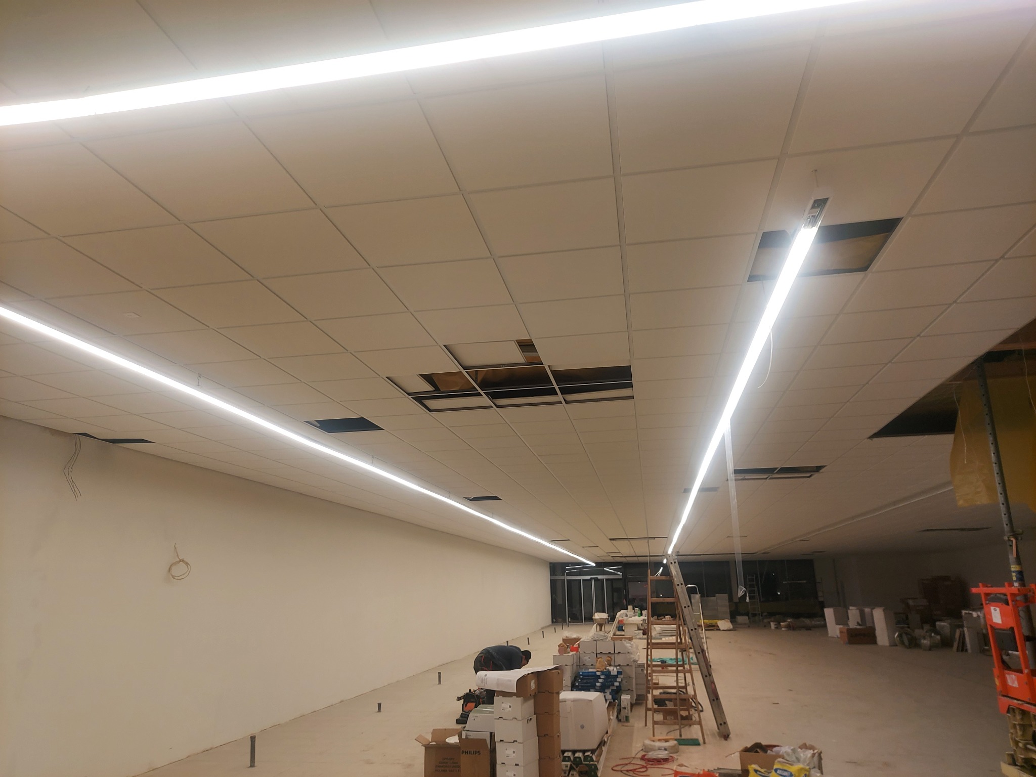 Wnętrze komercyjne w trakcie montażu oświetlenia liniowego LED w suficie podwieszanym, widoczne braki w panelach sufitowych i rozłożone materiały instalacyjne na podłodze.