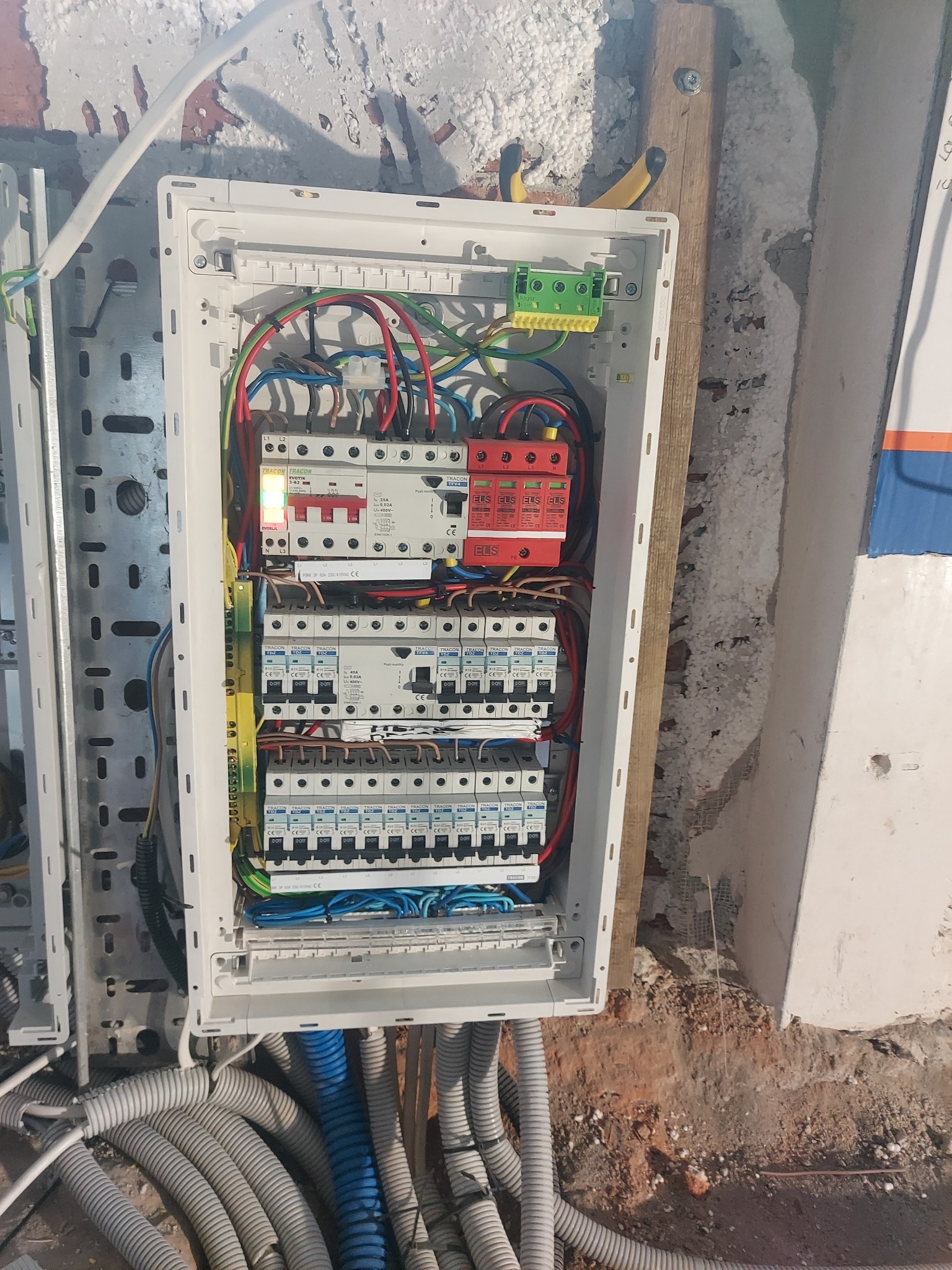 Rozdzielnica elektryczna z zainstalowanymi bezpiecznikami, wyłącznikami i przewodami w kolorach niebieskim, czerwonym, zielonym i żółtym, zamontowana na ścianie z widocznymi śladami prac...