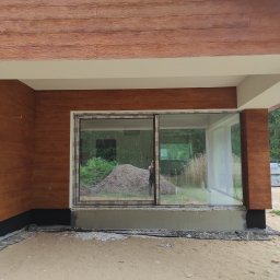 Mariusz Jurczak KURP-BUD - Elewacja budynku w trakcie wykończenia z drewnopodobnymi panelami i dużymi przesuwnymi oknami, widoczny fragment izolacji fundamentów i przygotowane podłoże.