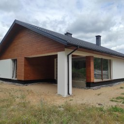 Mariusz Jurczak KURP-BUD - Nowoczesny dom jednorodzinny z drewnianą elewacją i białymi ścianami, czarnym dachem i kominem, widok z zewnątrz na etapie wykończenia terenu.