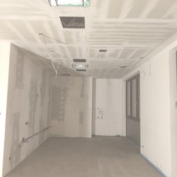 Mariusz Jurczak KURP-BUD - Wnętrze w trakcie prac wykończeniowych: surowa ściana z widocznymi łączeniami, podwieszany sufit z perforacją i otworami na instalacje, przygotowane do dalszych prac.