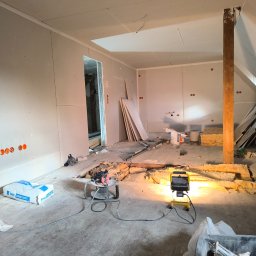 Mariusz Jurczak KURP-BUD - Poddasze w trakcie remontu: ściany wykończone płytami gipsowo-kartonowymi, widoczne narzędzia, materiały budowlane, oświetlenie robocze i częściowo odsłonięta izolacja podłogi.