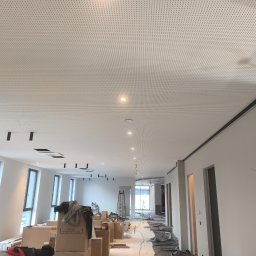 Mariusz Jurczak KURP-BUD - Montaż sufitu podwieszanego z perforowanej płyty w przestrzeni biurowej w trakcie wykończenia, widoczne kable i oprawy oświetleniowe, kartony z materiałami budowlanymi na podłodze.