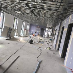 Mariusz Jurczak KURP-BUD - Wnętrze w trakcie montażu płyt gipsowo-kartonowych, widoczna metalowa konstrukcja sufitu podwieszanego, ściany przygotowane pod gładzie, drabiny, narzędzia i materiały budowlane porozrzucane...