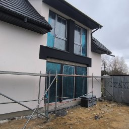 Mariusz Jurczak KURP-BUD - Elewacja domu w trakcie prac: biała ściana, czarne obramowania okien zabezpieczonych folią, rusztowanie, farby. Widok z boku, pochmurne niebo.
