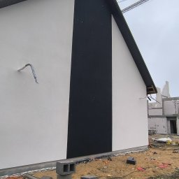 Mariusz Jurczak KURP-BUD - Nowoczesna elewacja domu jednorodzinnego w kolorze białym i czarnym, z widocznymi elementami instalacji i placem budowy w tle. Minimalistyczny design.