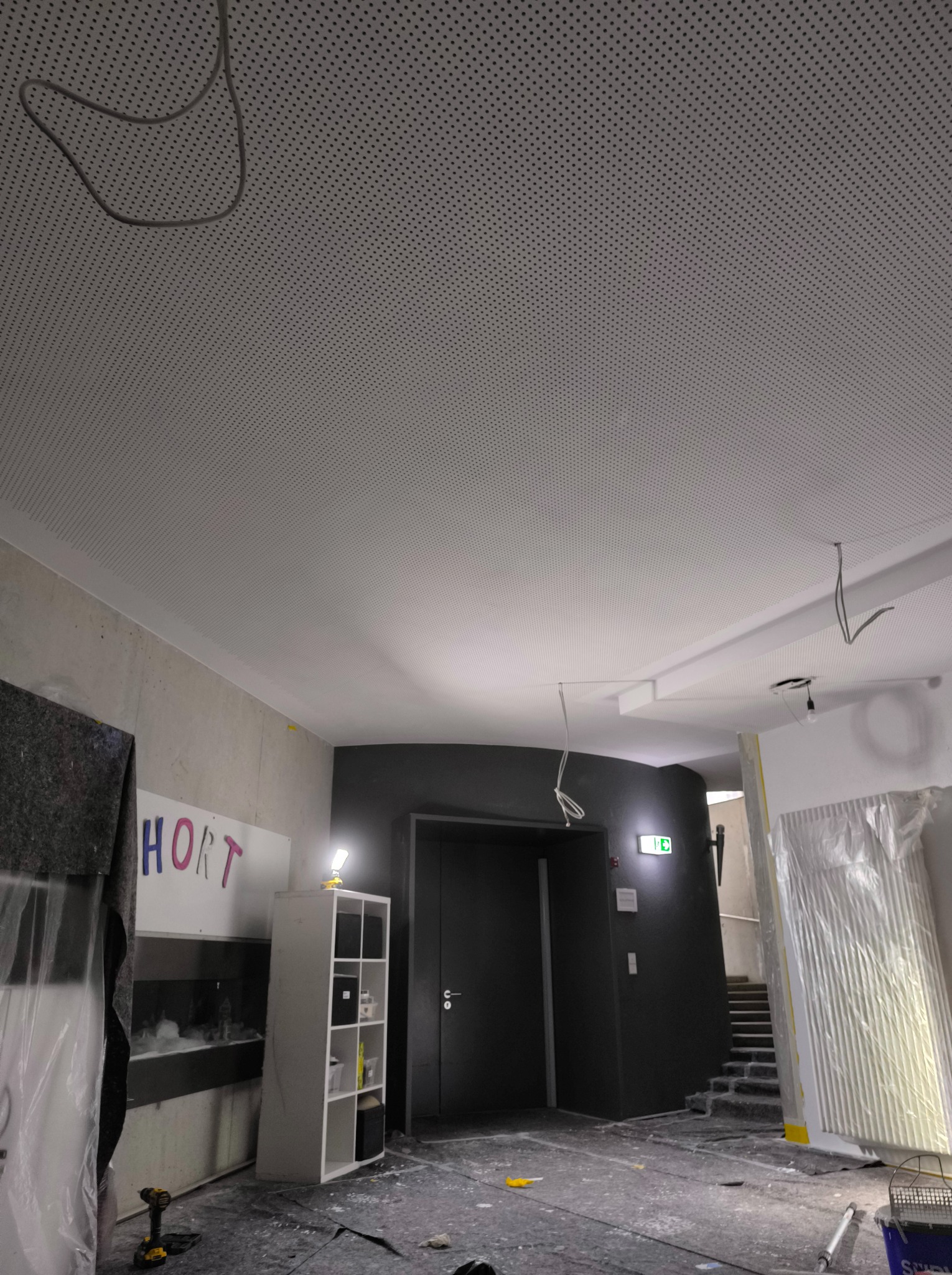 Perforowany sufit podwieszany w trakcie montażu, widoczne kable, zabezpieczone folią elementy wnętrza i narzędzia na podłodze.