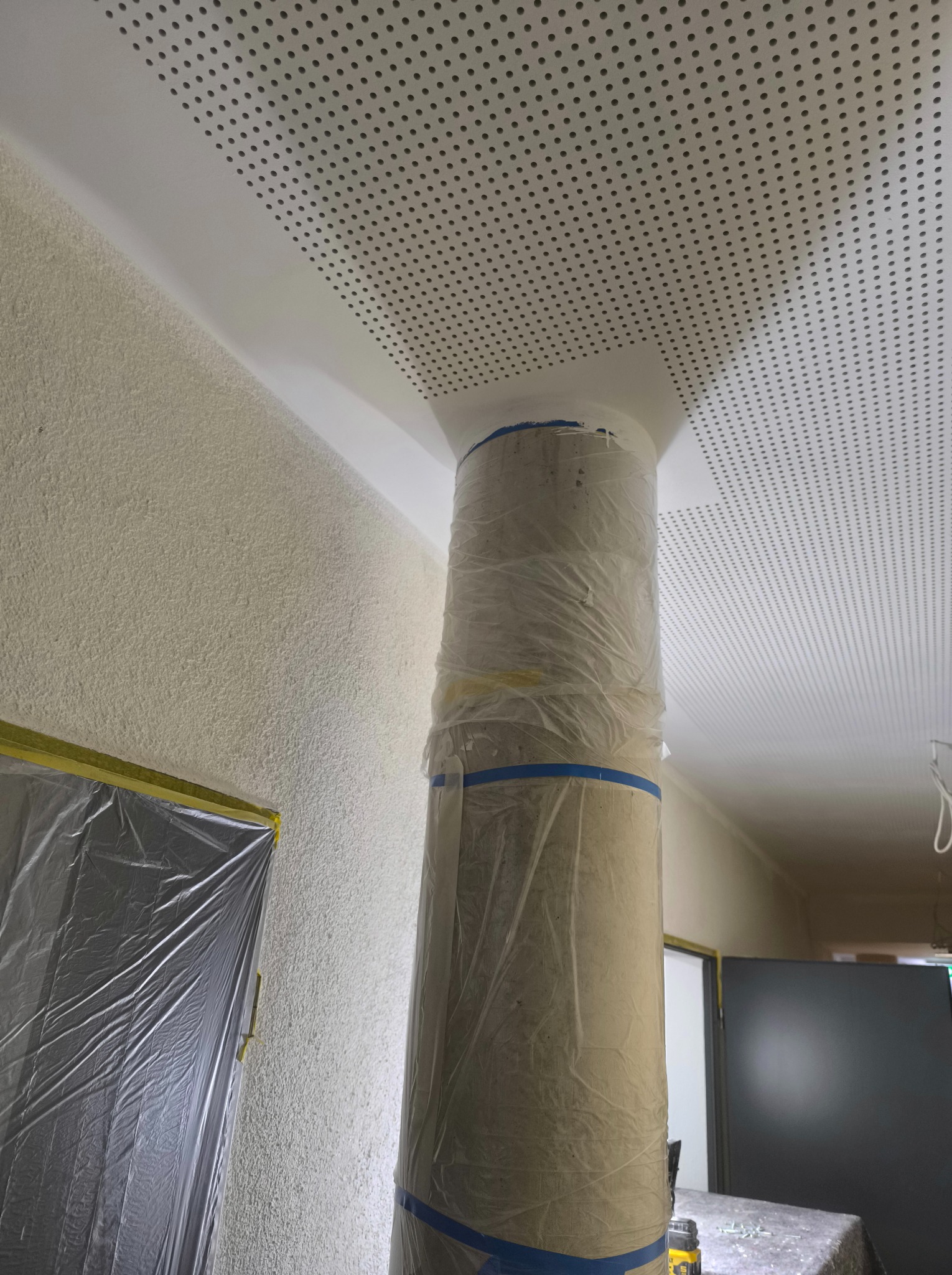 Przygotowanie do malowania: kolumna zabezpieczona folią i taśmą malarską, ściana z fakturą i sufit z perforacją, okno zakryte folią ochronną.