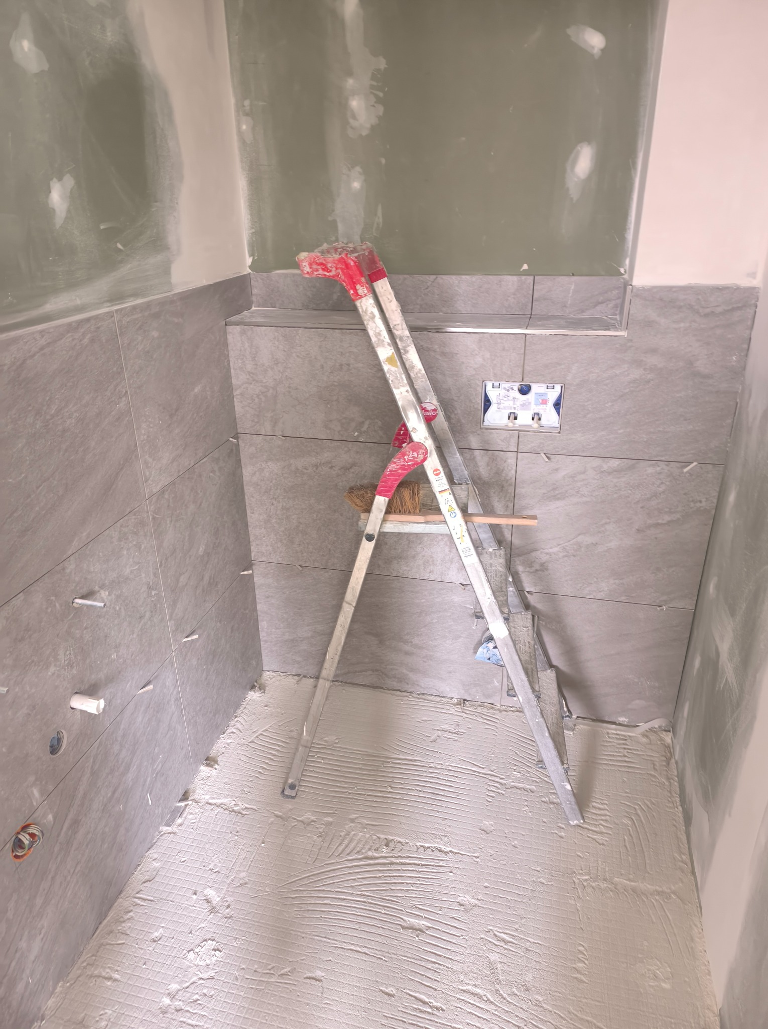Wnętrze łazienki w trakcie remontu: szare płytki na ścianach, rozprowadzona zaprawa na podłodze, drabina oparta o ścianę z wystającymi elementami instalacji wodnej.