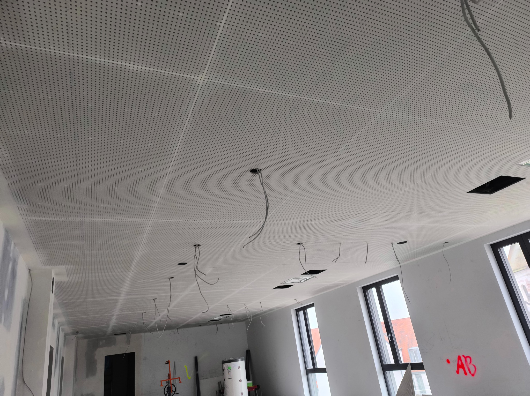 Sufit podwieszany z perforowanej płyty w trakcie montażu, widoczne wystające kable instalacji elektrycznej i ślady prac wykończeniowych na ścianach.