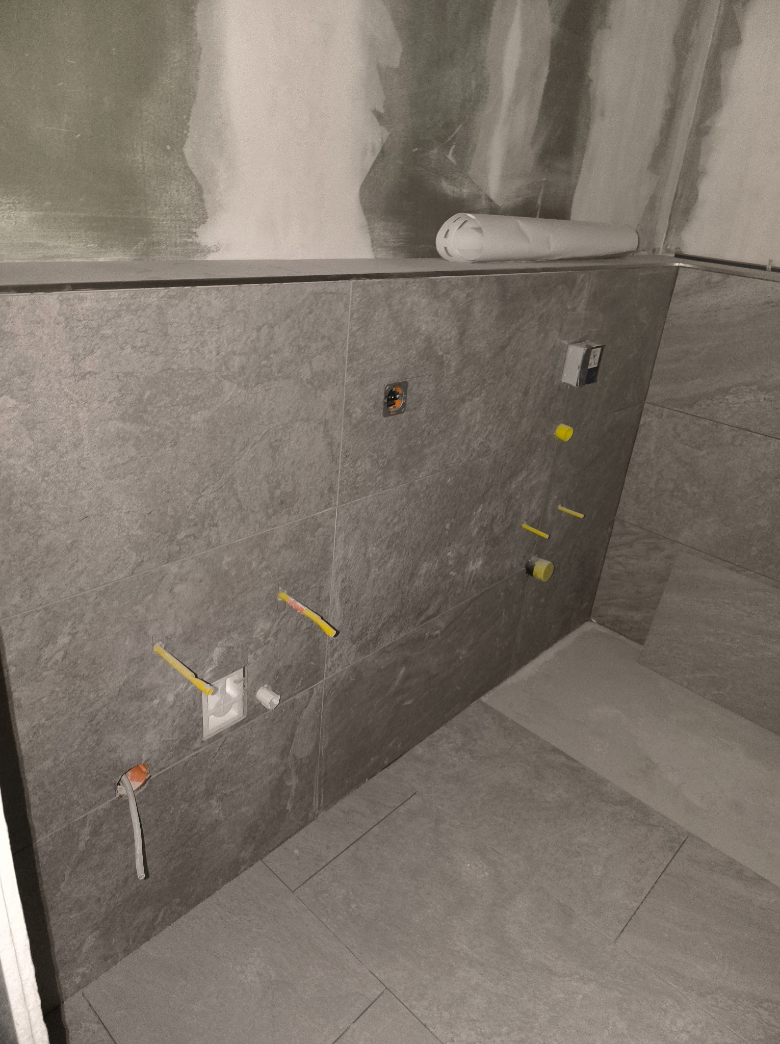 Szare płytki ścienne i podłogowe w trakcie montażu w łazience, widoczne wystające rury i kable, ściany częściowo pokryte płytą kartonowo-gipsową, rolka papieru zabezpieczającego na górnej krawędzi...