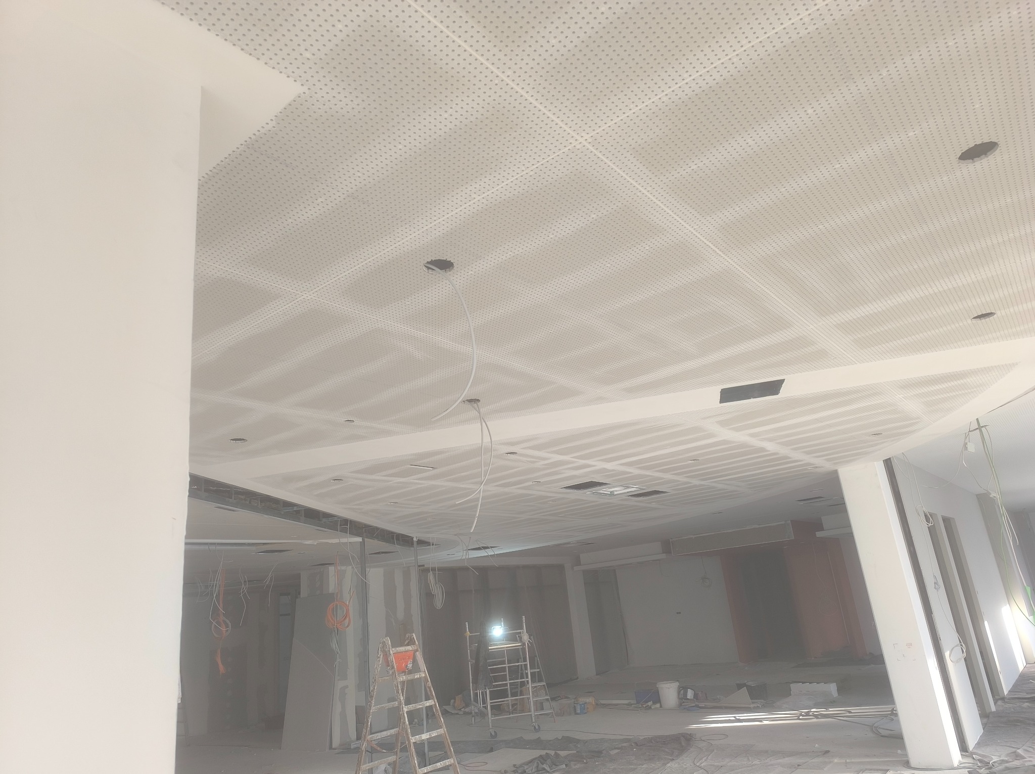 Montaż sufitu podwieszanego z płyt perforowanych w pomieszczeniu komercyjnym, widoczne rusztowania i przewody elektryczne.