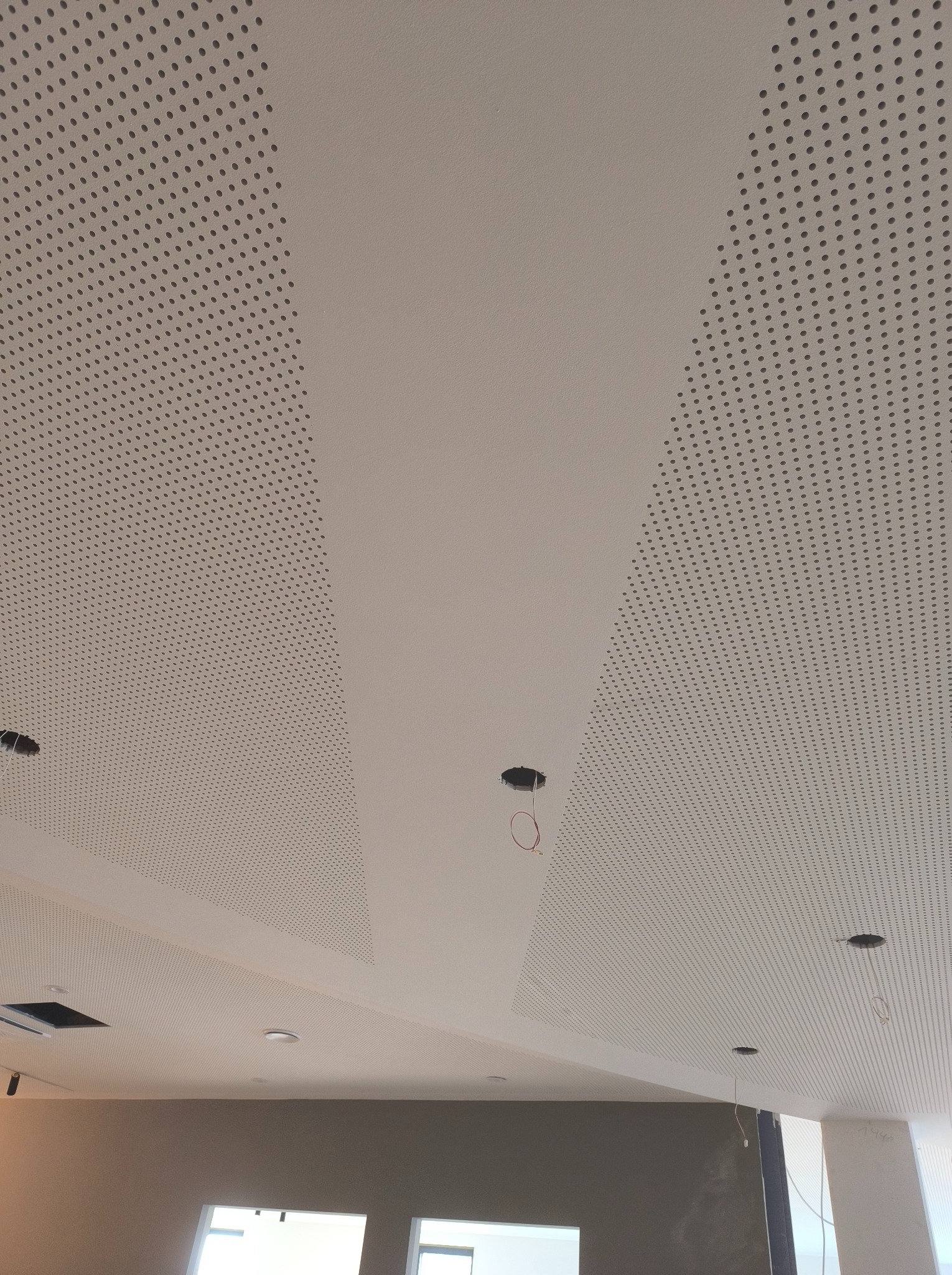 Sufit podwieszany z perforowanymi panelami i gładkimi pasami, widoczne otwory na oświetlenie i wystające przewody.