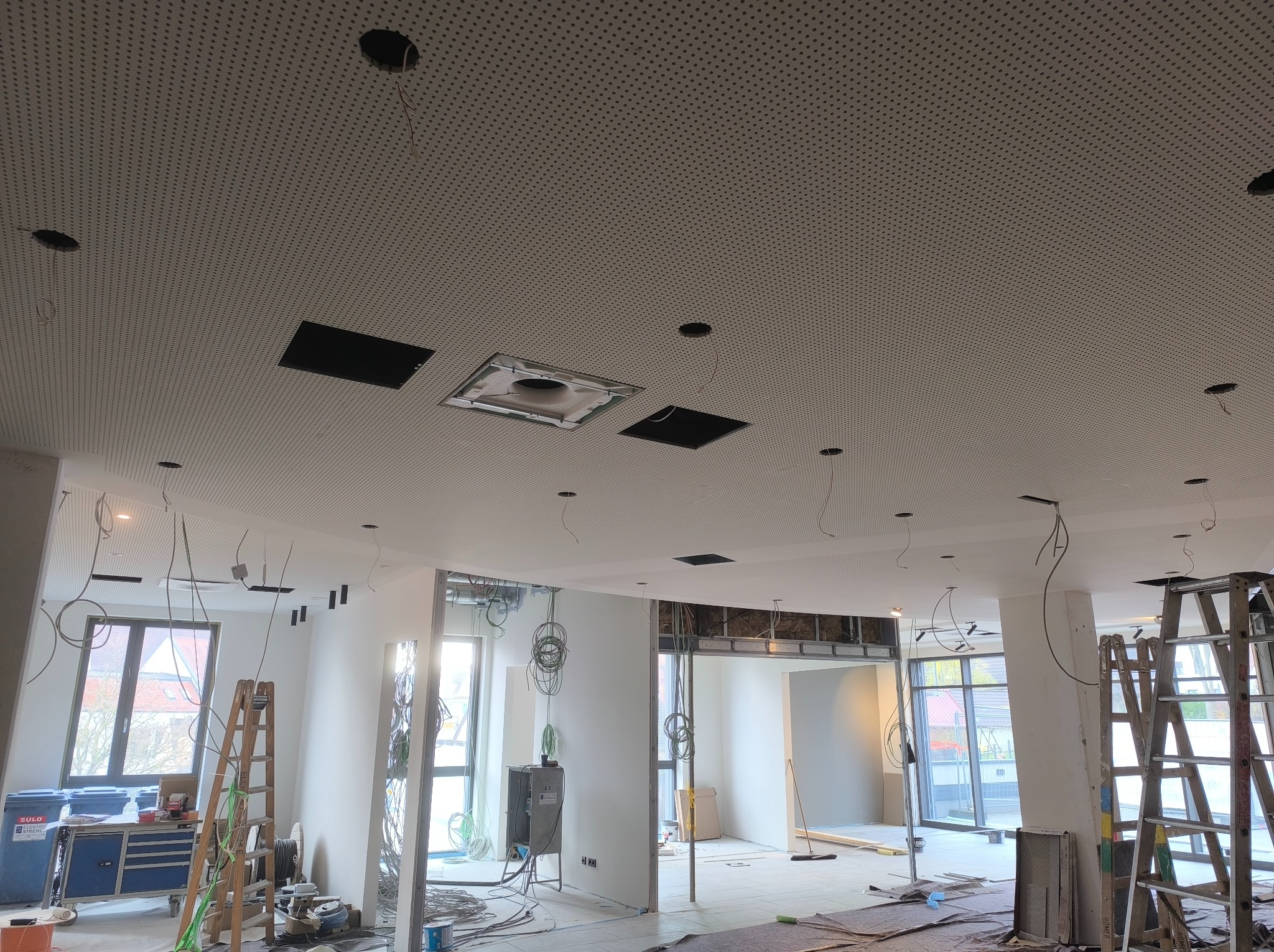 Widok wnętrza w trakcie prac wykończeniowych, z widocznym perforowanym sufitem podwieszanym z otworami na oświetlenie i instalacje, częściowo zamontowane ściany działowe, oraz drabiny i narzędzia...