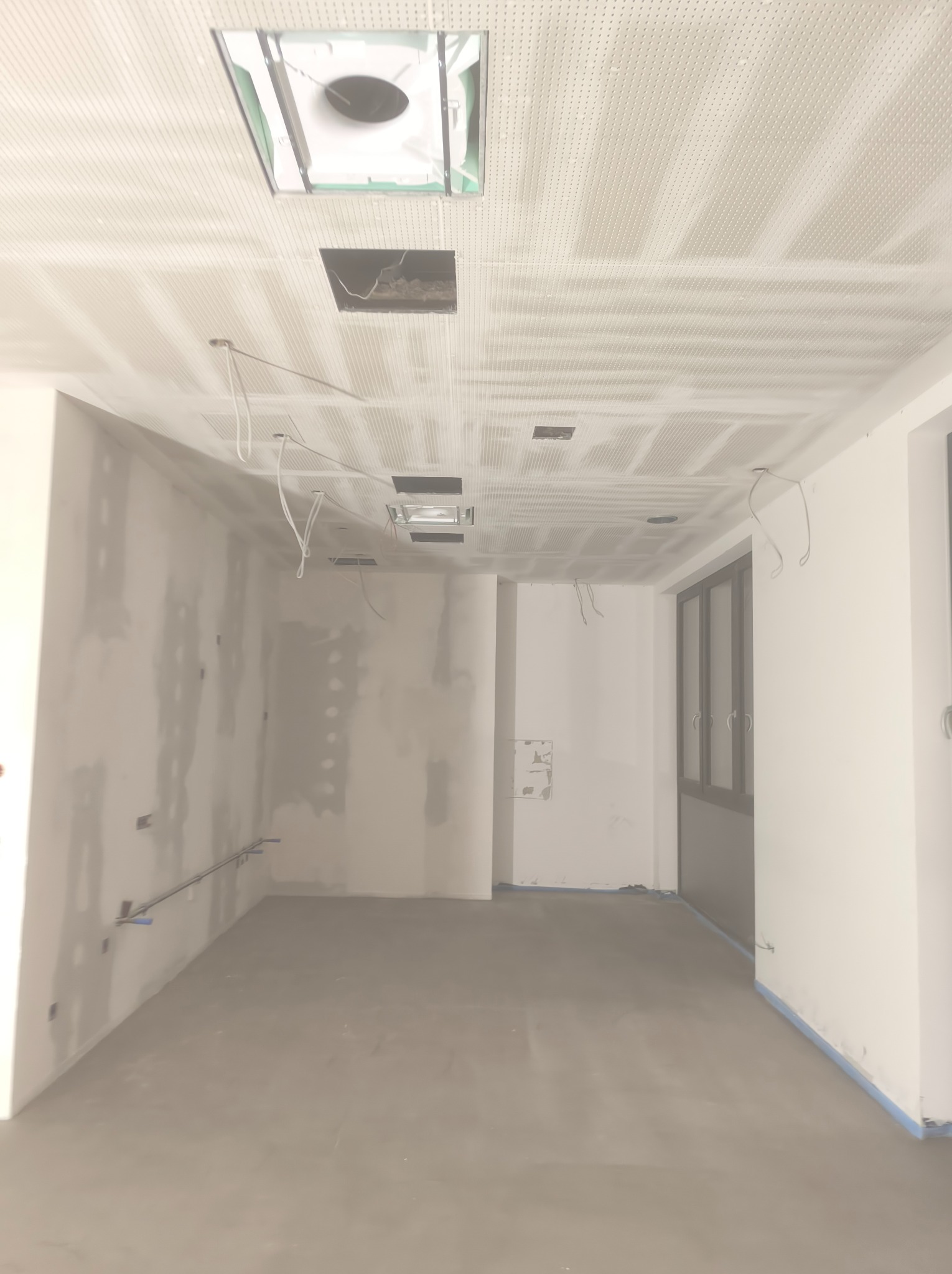Wnętrze w trakcie prac wykończeniowych: surowa ściana z widocznymi łączeniami, podwieszany sufit z perforacją i otworami na instalacje, przygotowane do dalszych prac.