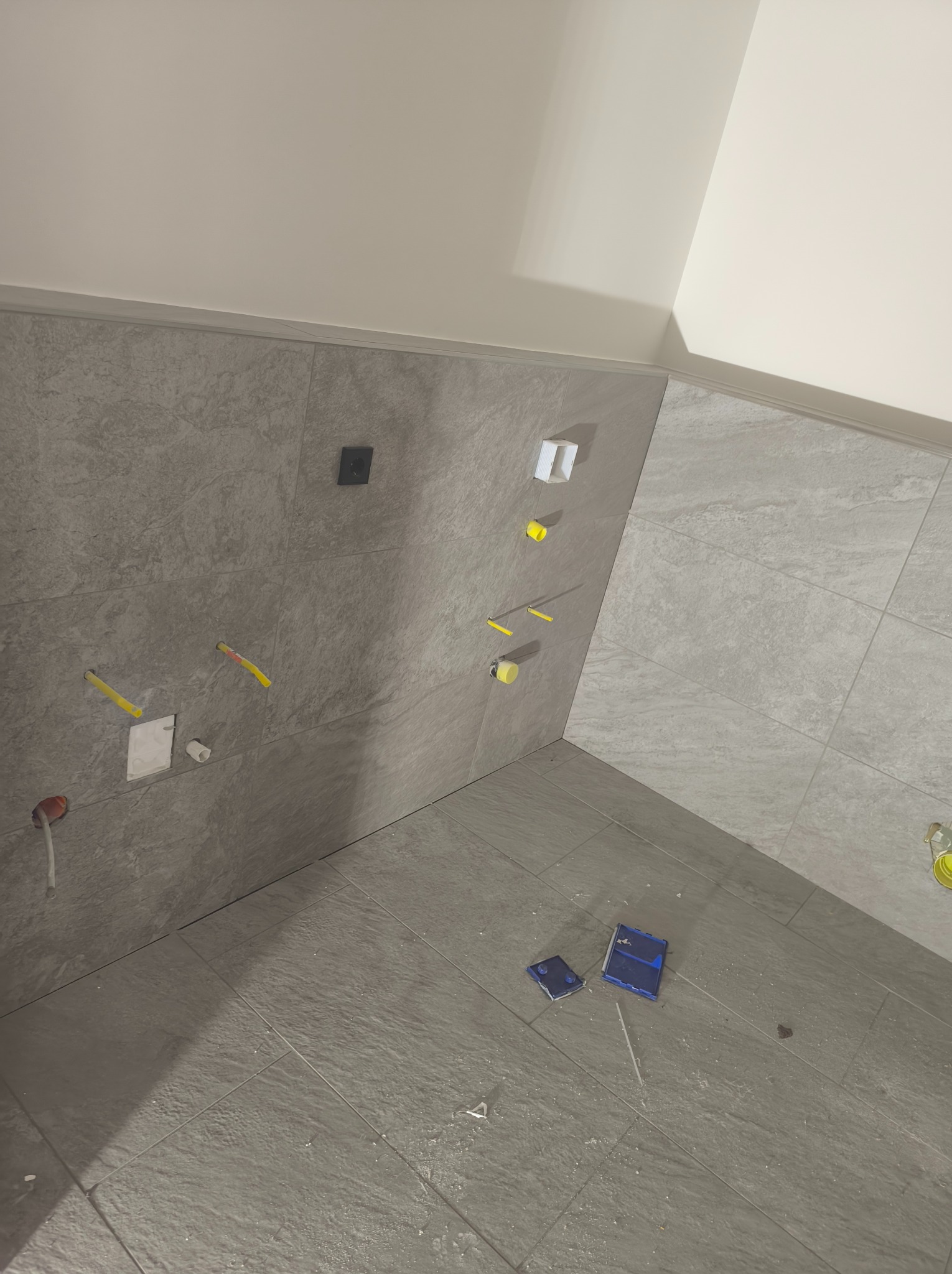 Szare płytki ceramiczne na ścianach i podłodze w łazience w trakcie remontu, widoczne wystające kable elektryczne i narzędzia glazurnicze na podłodze.