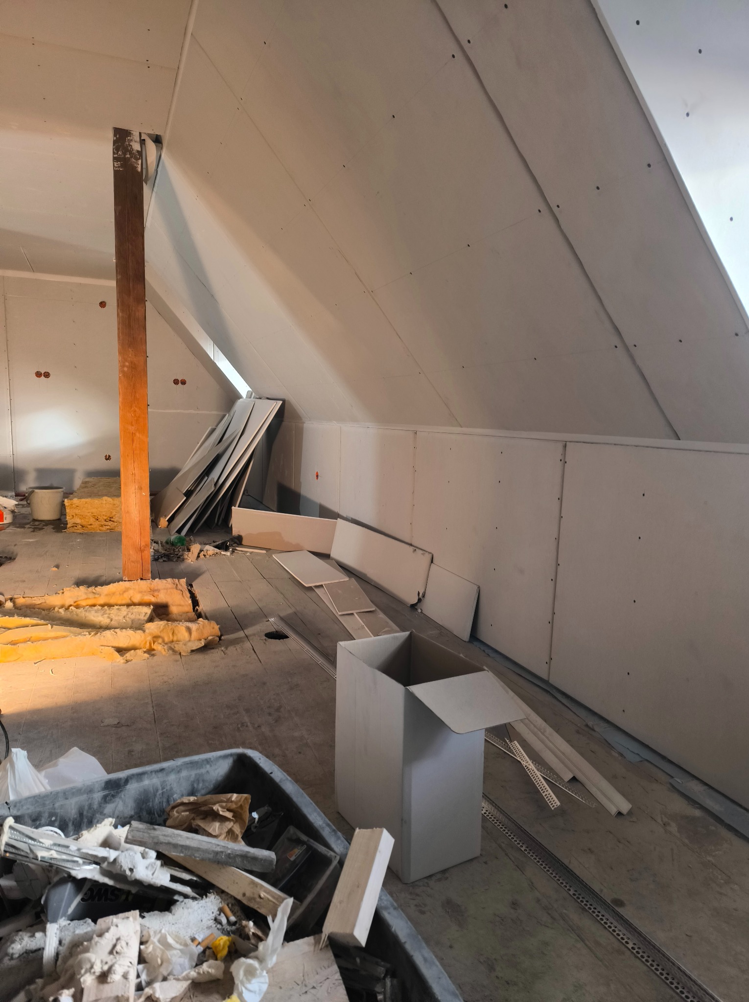 Poddasze w trakcie wykańczania płytami kartonowo-gipsowymi, widoczne płyty na ścianach i skosach, izolacja, drewniany słup konstrukcyjny oraz pojemnik z odpadami budowlanymi na drewnianej podłodze.