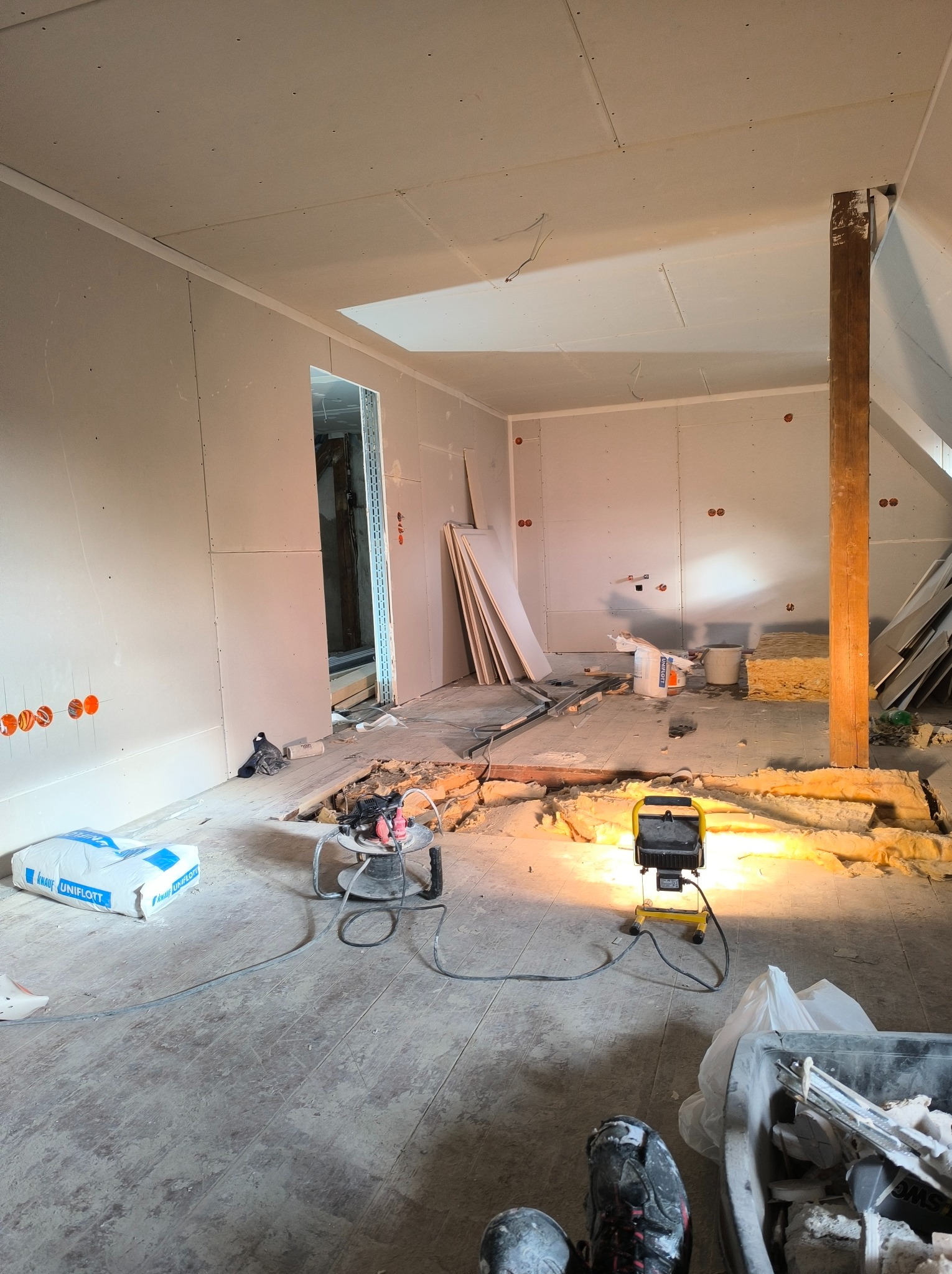 Poddasze w trakcie remontu: ściany wykończone płytami gipsowo-kartonowymi, widoczne narzędzia, materiały budowlane, oświetlenie robocze i częściowo odsłonięta izolacja podłogi.