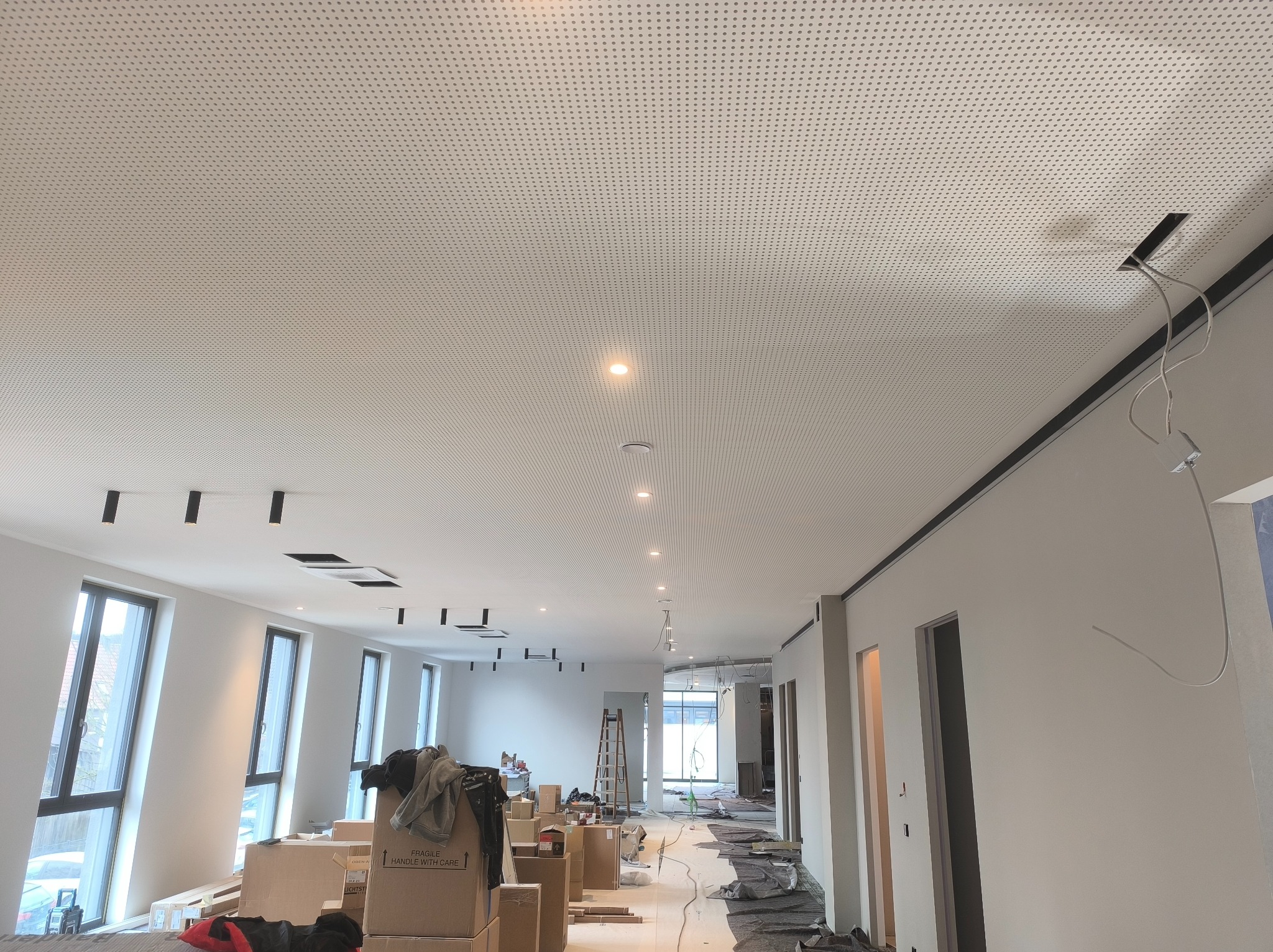 Montaż sufitu podwieszanego z perforowanej płyty w przestrzeni biurowej w trakcie wykończenia, widoczne kable i oprawy oświetleniowe, kartony z materiałami budowlanymi na podłodze.