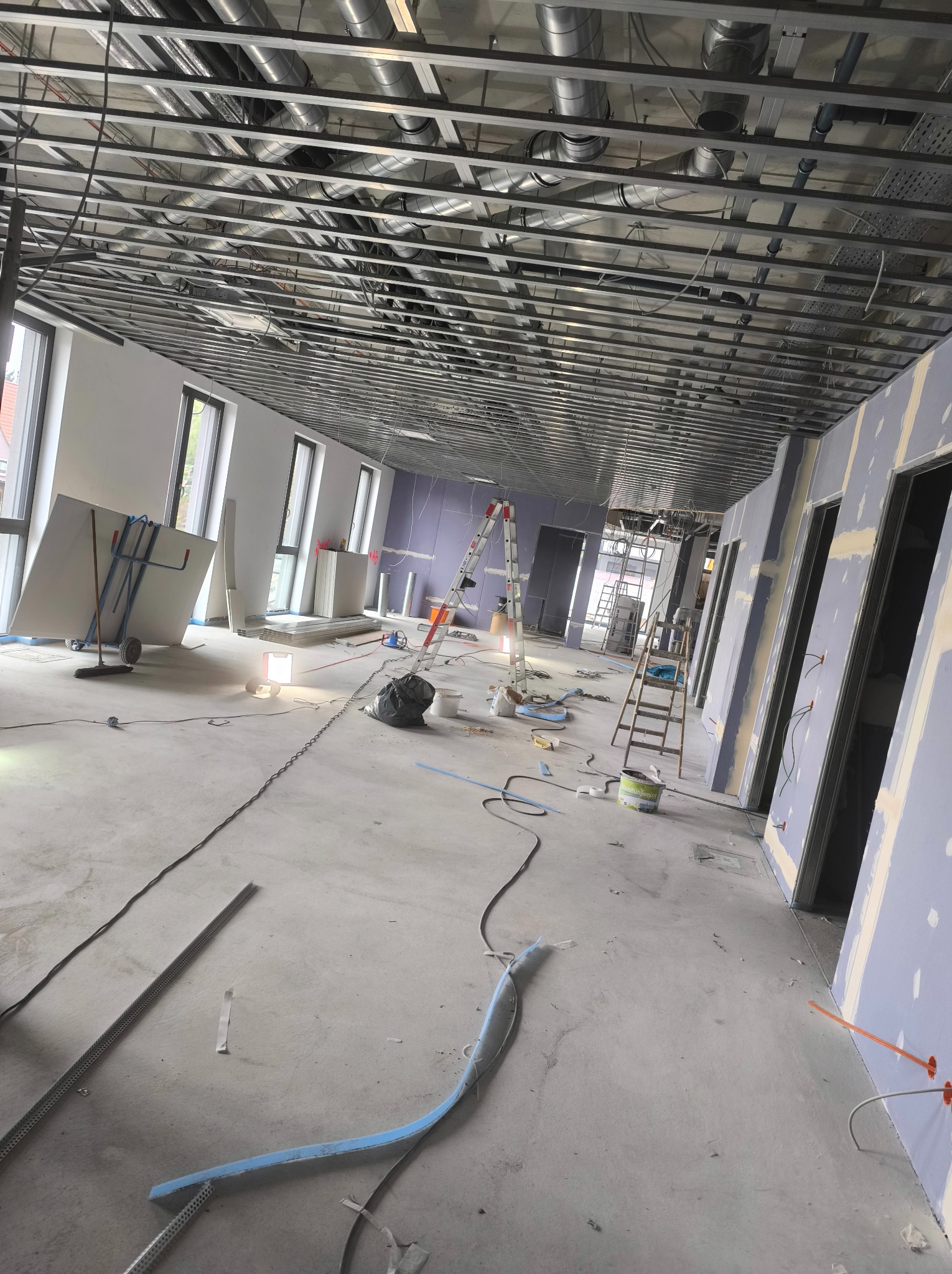 Wnętrze w trakcie montażu płyt gipsowo-kartonowych, widoczna metalowa konstrukcja sufitu podwieszanego, ściany przygotowane pod gładzie, drabiny, narzędzia i materiały budowlane porozrzucane...