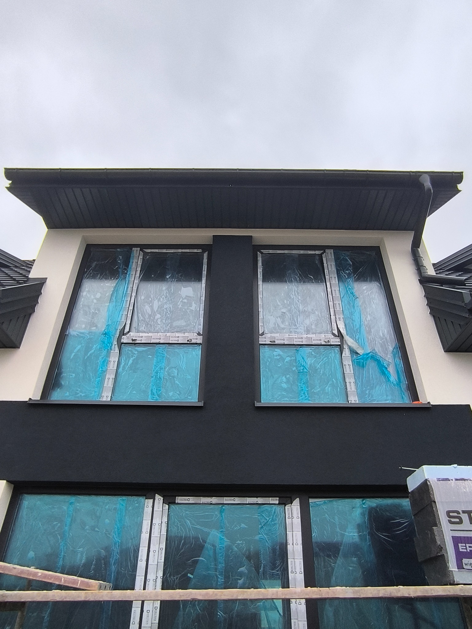 Fragment elewacji domu z zabezpieczonymi folią oknami, przygotowany do prac wykończeniowych. Widoczne rusztowanie i materiały budowlane.