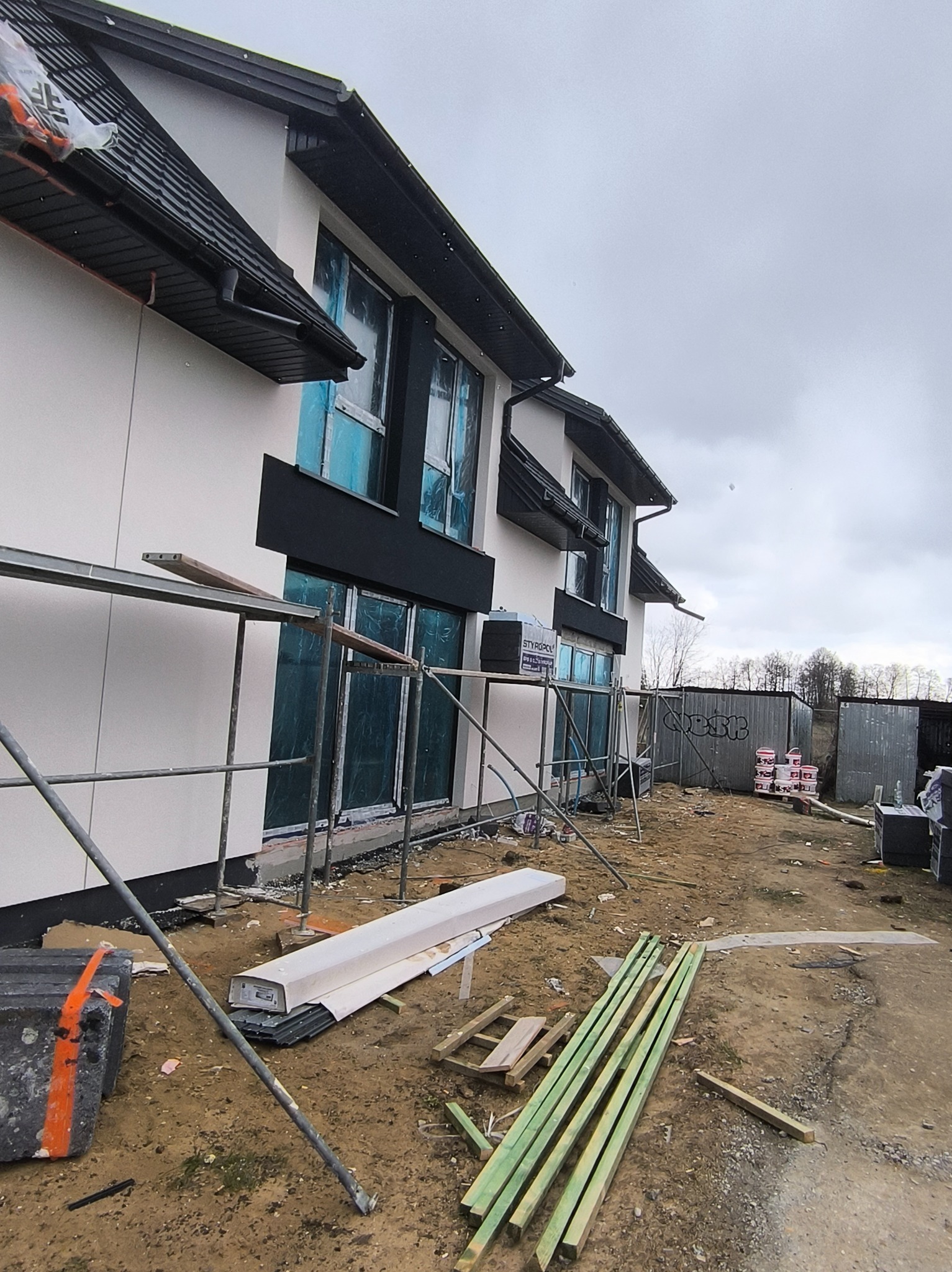 Ocieplona elewacja domu w trakcie wykończenia, z widocznymi rusztowaniami i materiałami budowlanymi. Okna zabezpieczone folią. Widoczne styropiany.