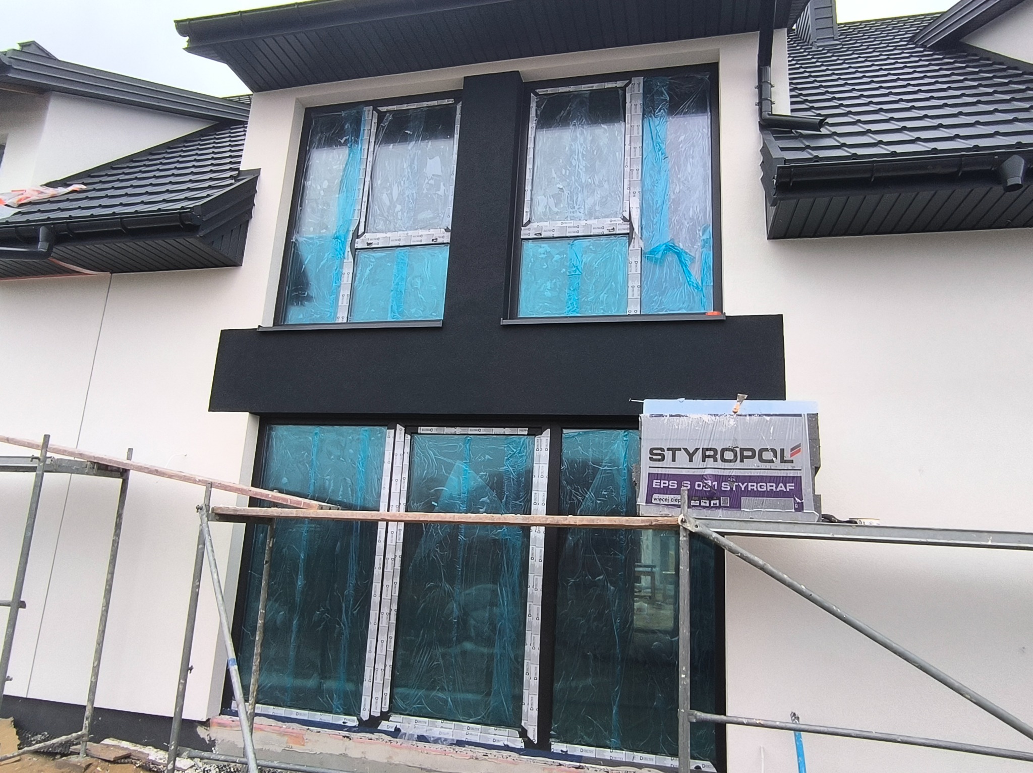 Elewacja domu w trakcie ocieplania, okna zabezpieczone folią, widoczne rusztowanie i paczka styropianu STYROPOL EPS S 031. Biała ściana i czarne elementy wykończeniowe.