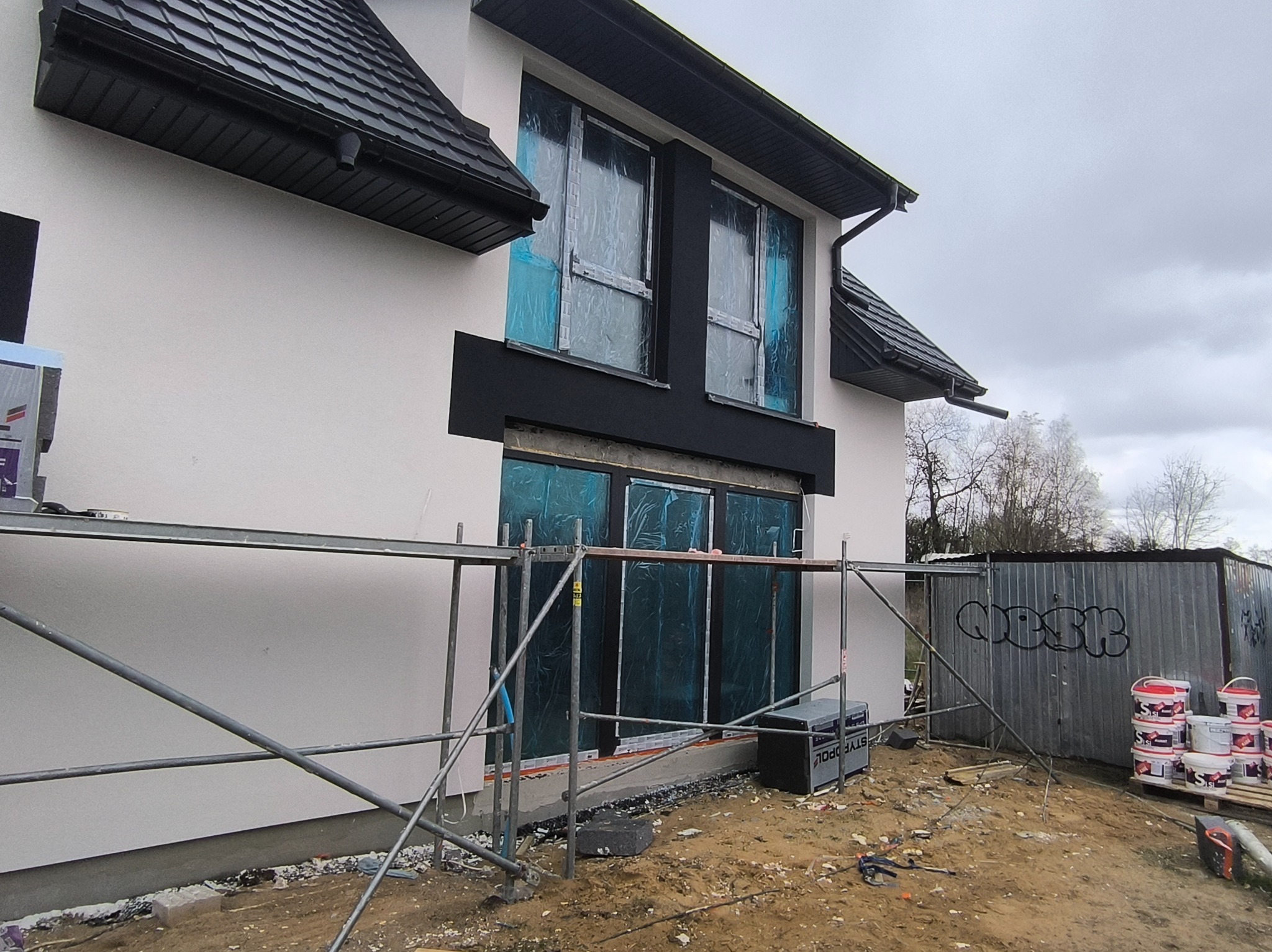 Elewacja domu w trakcie prac: biała ściana, czarne obramowania okien zabezpieczonych folią, rusztowanie, farby. Widok z boku, pochmurne niebo.