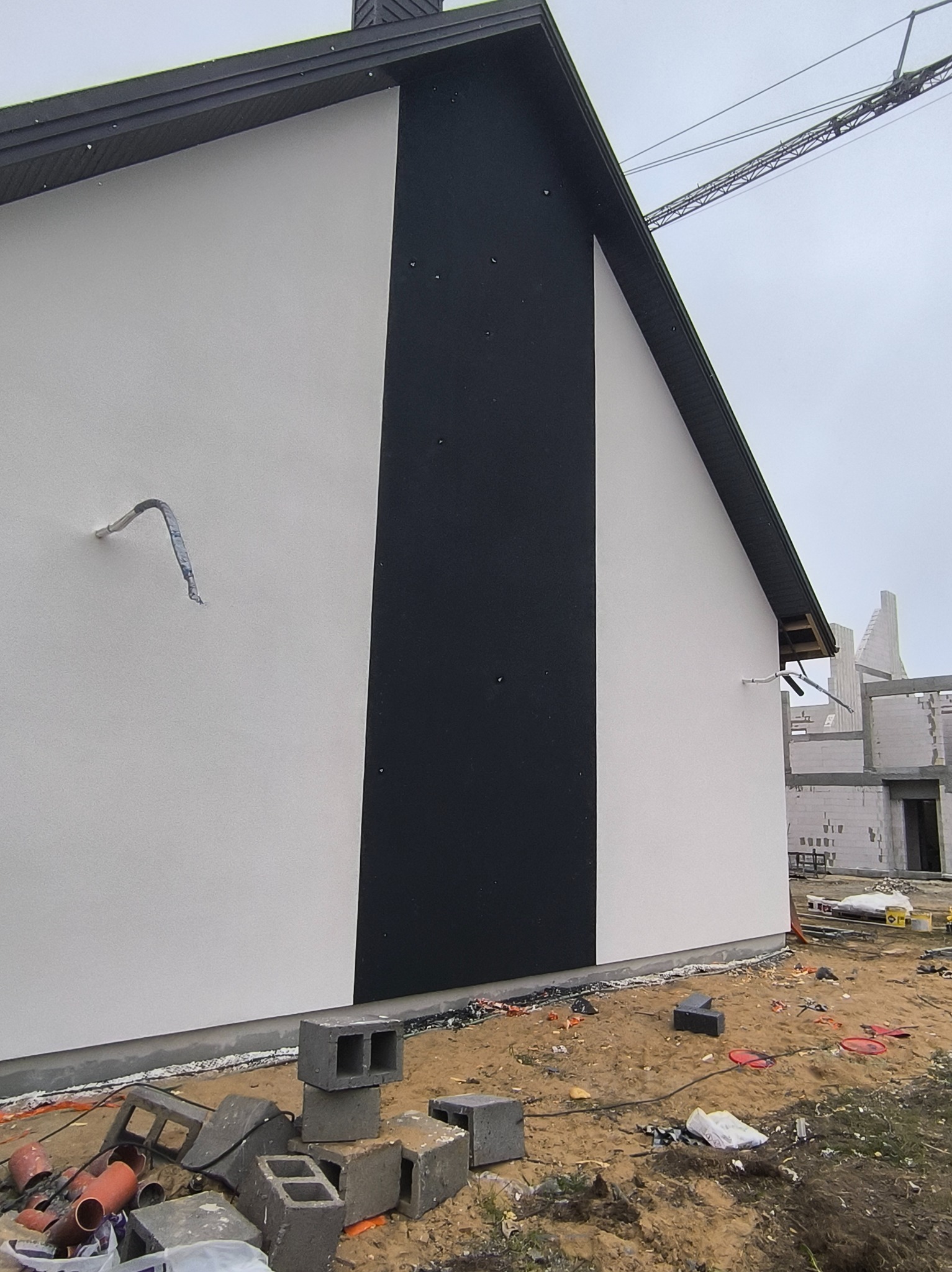 Nowoczesna elewacja domu jednorodzinnego w kolorze białym i czarnym, z widocznymi elementami instalacji i placem budowy w tle. Minimalistyczny design.