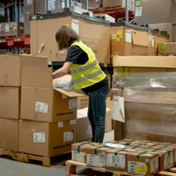 Pracownica w żółtej kamizelce kompletuje zamówienie w magazynie, otwierając karton z napisem 'ASSEMBLED IN MEXICO' na tle regałów z paletami i opakowanymi towarami.