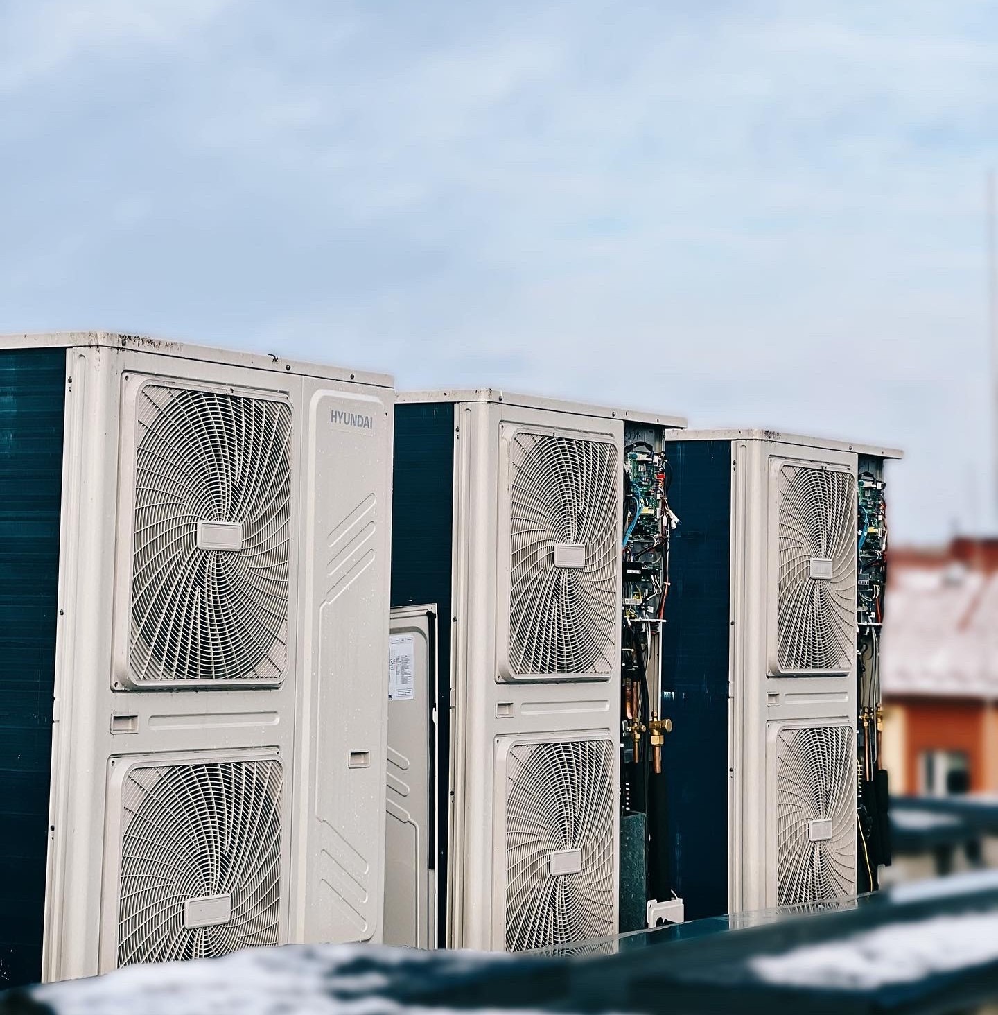 Rząd zewnętrznych jednostek klimatyzacyjnych marki Hyundai na dachu, z otwartą jedną obudową ukazującą wewnętrzne okablowanie i rury, na tle błękitnego nieba.