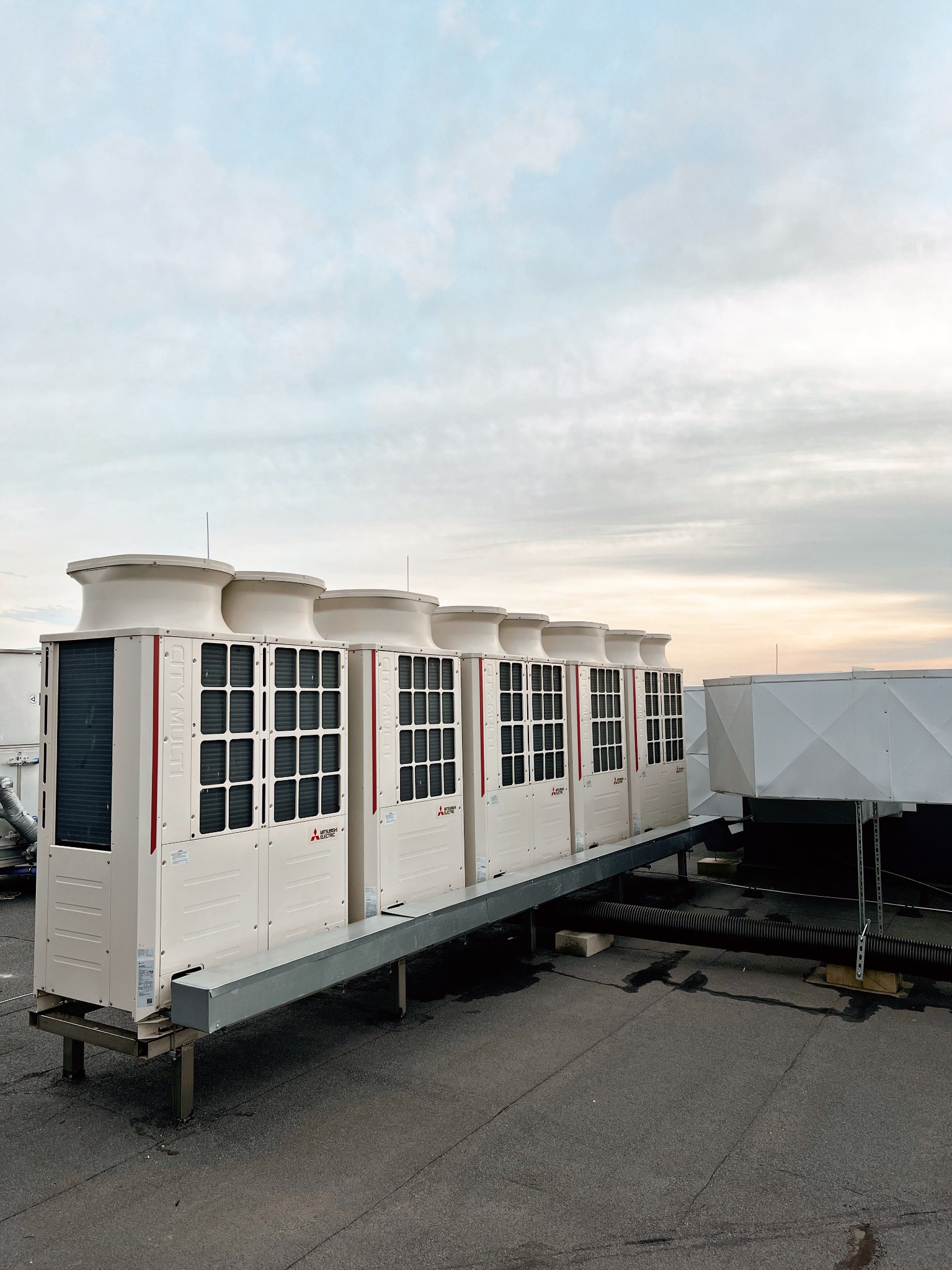 Rząd kremowych, modułowych klimatyzatorów przemysłowych Mitsubishi Electric ustawionych na metalowej konstrukcji na płaskim dachu, z widocznymi przewodami i elementami wentylacyjnymi, na tle...