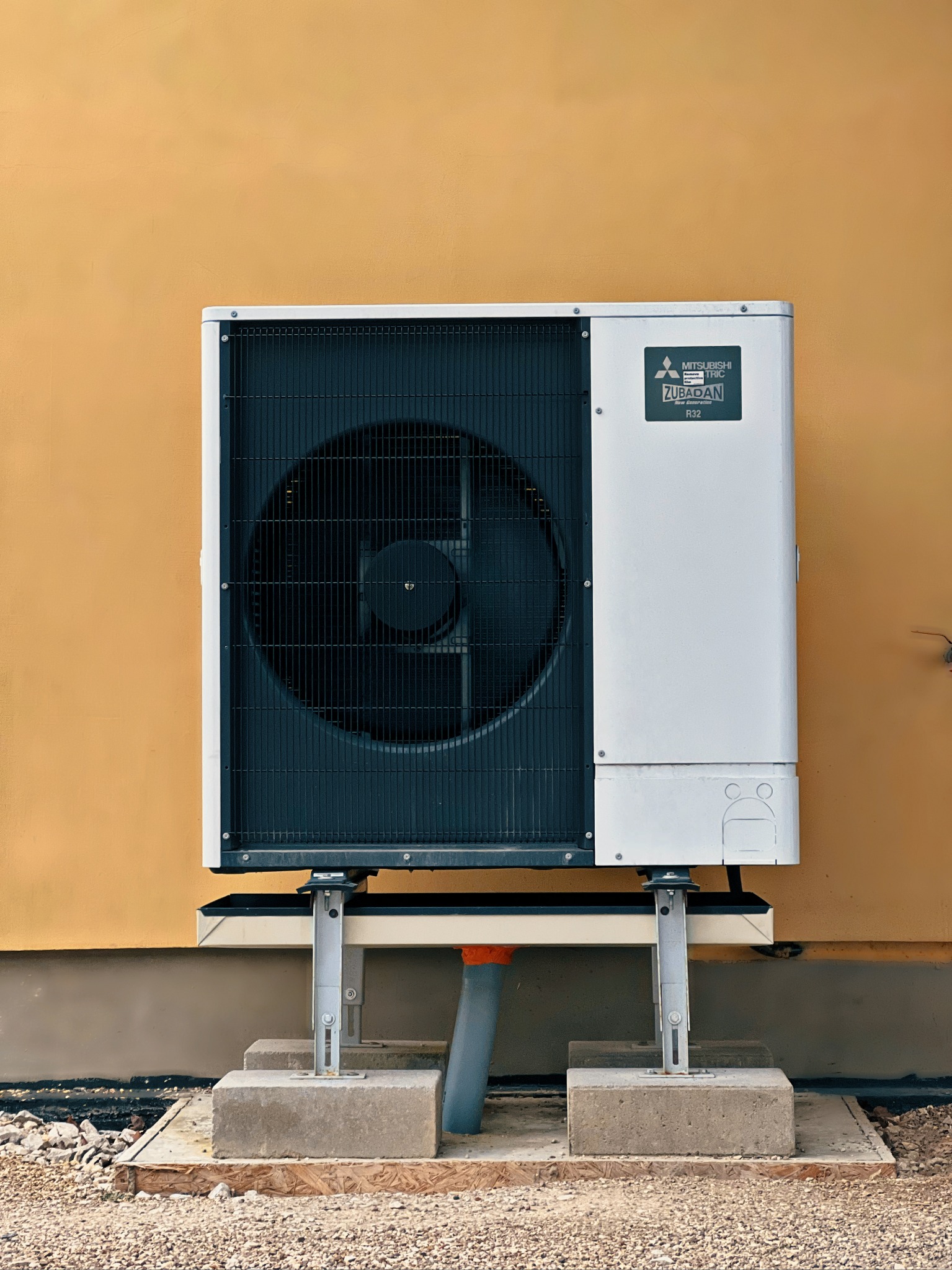 Biała jednostka zewnętrzna pompy ciepła Mitsubishi Zubadan R32 zamontowana na betonowych podstawach przy żółtej ścianie budynku. Widoczny szary odpływ kondensatu i żwirowe podłoże.