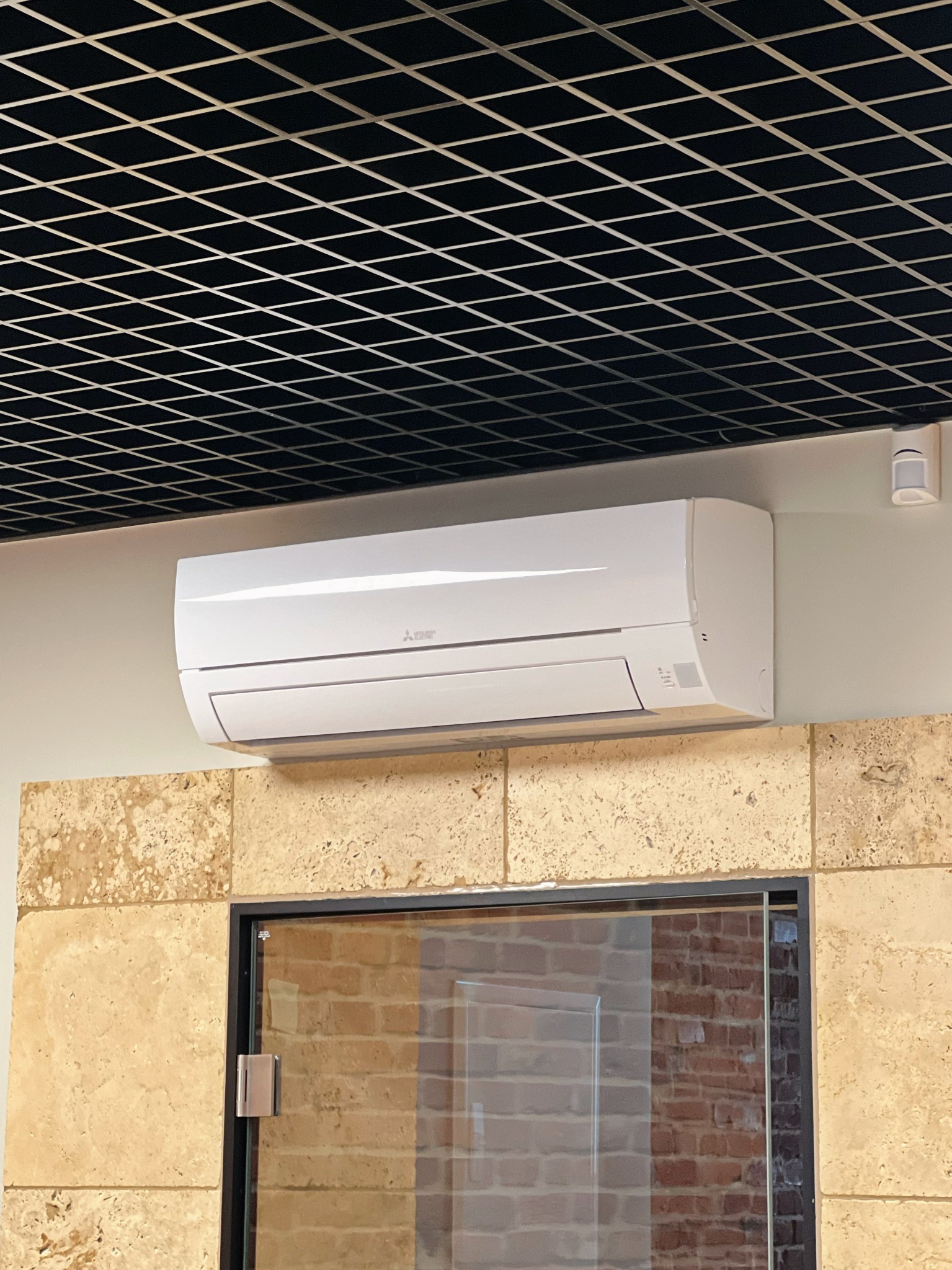 Biała jednostka klimatyzacyjna Mitsubishi zamontowana na ścianie z trawertynu w biurze, widoczna kratownica sufitowa i czujnik ruchu.