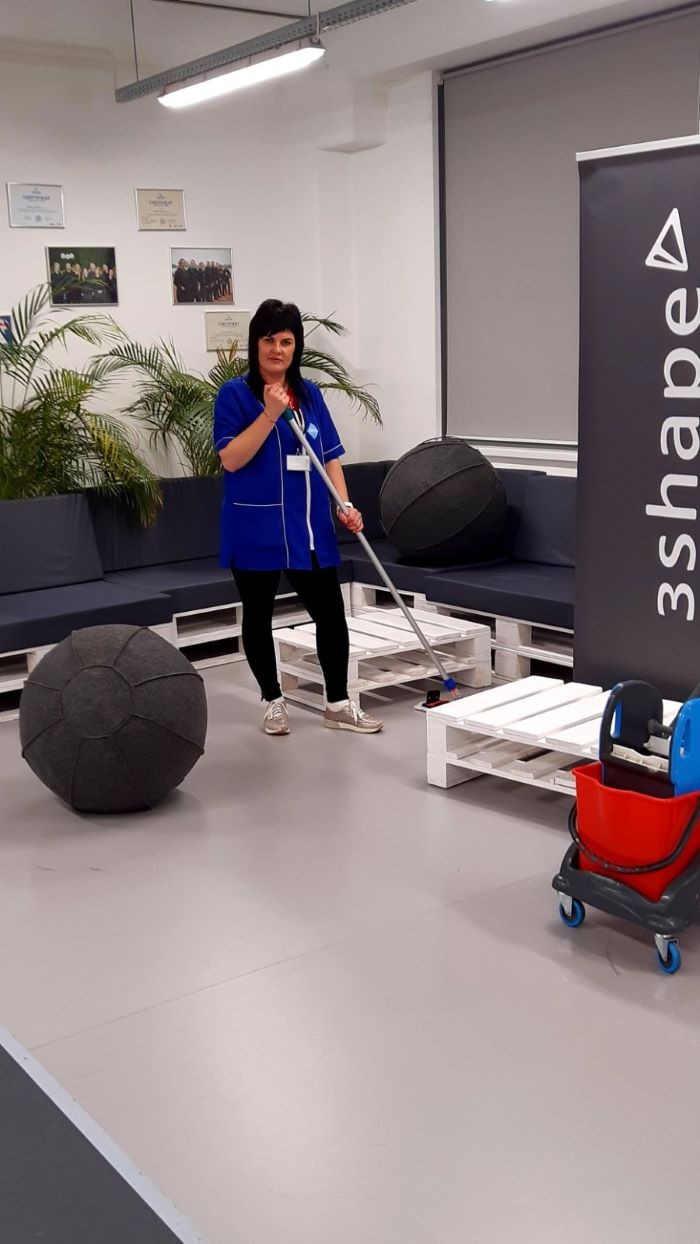 Pracownica w niebieskim uniformie myje podłogę mopem w nowoczesnym biurze z szarymi meblami z palet i dużymi kulistymi pufami.