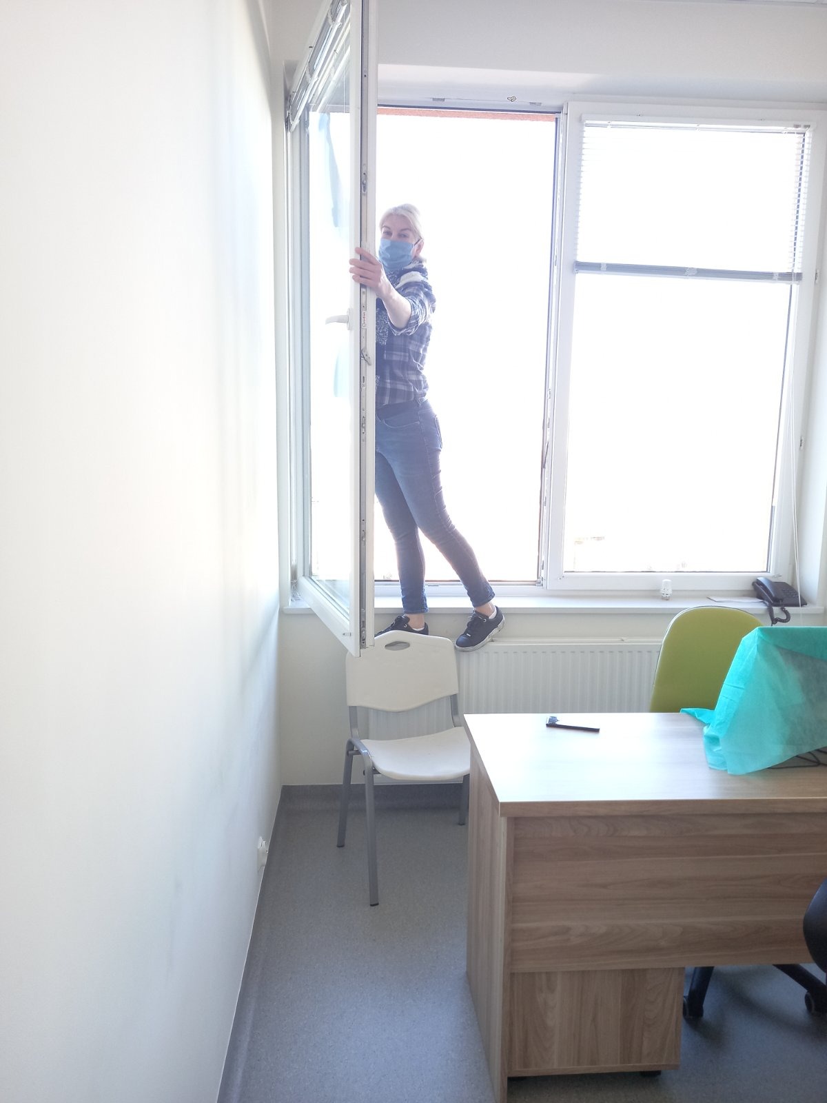 Kobieta w masce i kraciastej koszuli stoi na parapecie otwartego okna, trzymając ramę okienną, w jasnym biurze z biurkiem, krzesłami i kaloryferem.