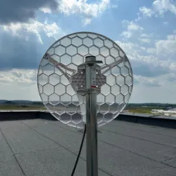 Antena telekomunikacyjna o strukturze plastra miodu zamontowana na metalowym słupie na płaskim dachu, z widokiem na odległy krajobraz i pochmurne niebo.