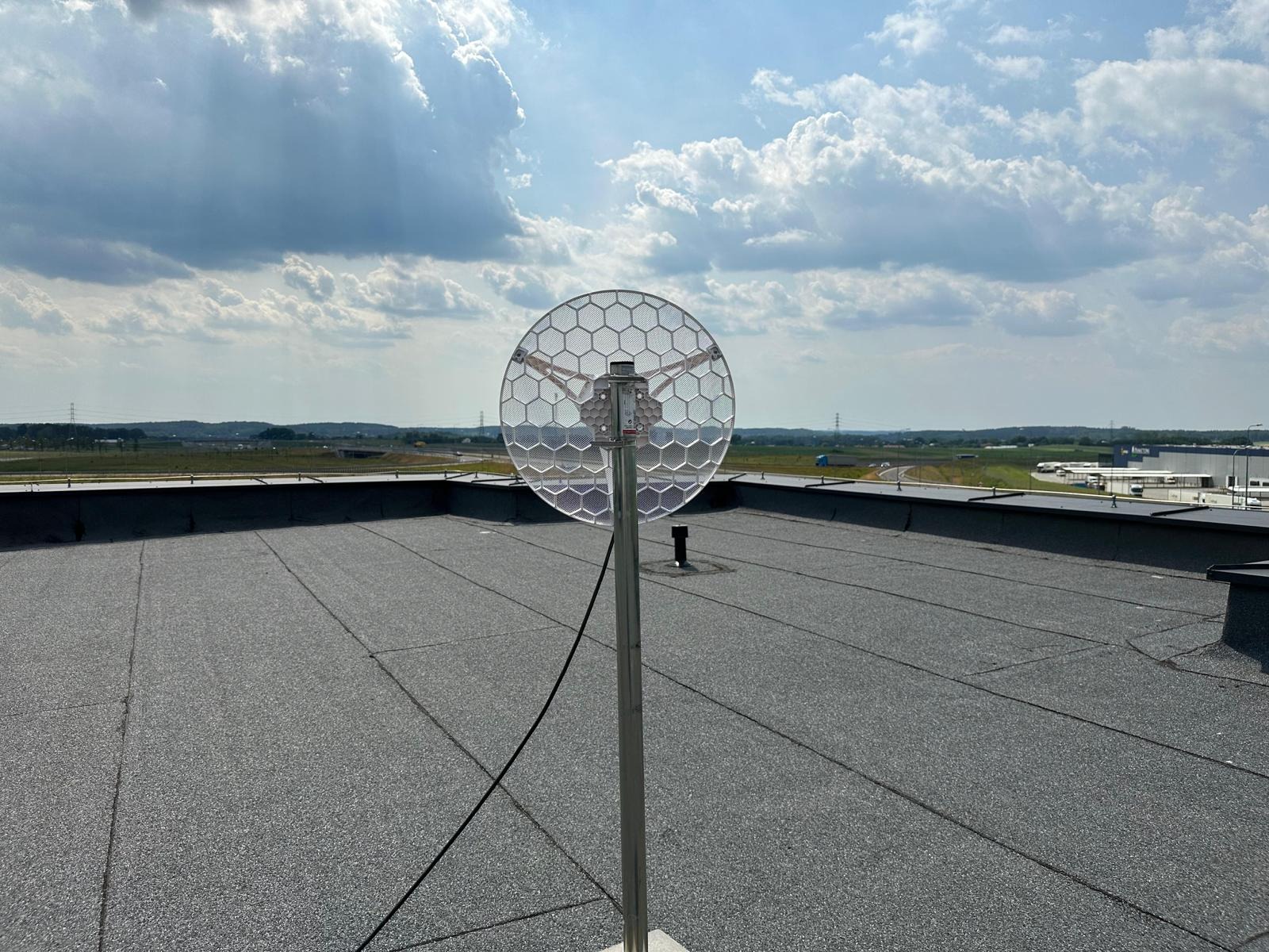 Antena telekomunikacyjna z siatką dyfrakcyjną zamontowana na płaskim dachu budynku przemysłowego, widok na odległe pola i autostradę pod błękitnym niebem z chmurami.