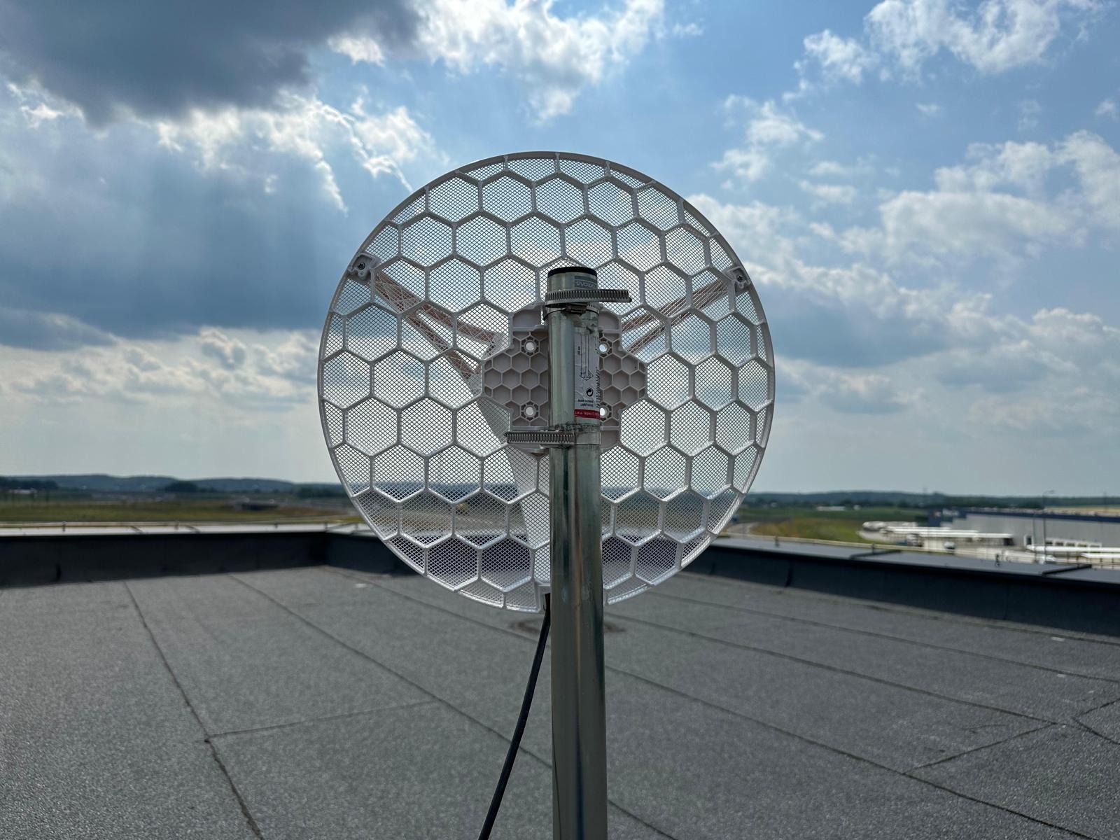 Antena telekomunikacyjna o strukturze plastra miodu zamontowana na metalowym słupie na płaskim dachu, z widokiem na odległy krajobraz i pochmurne niebo.