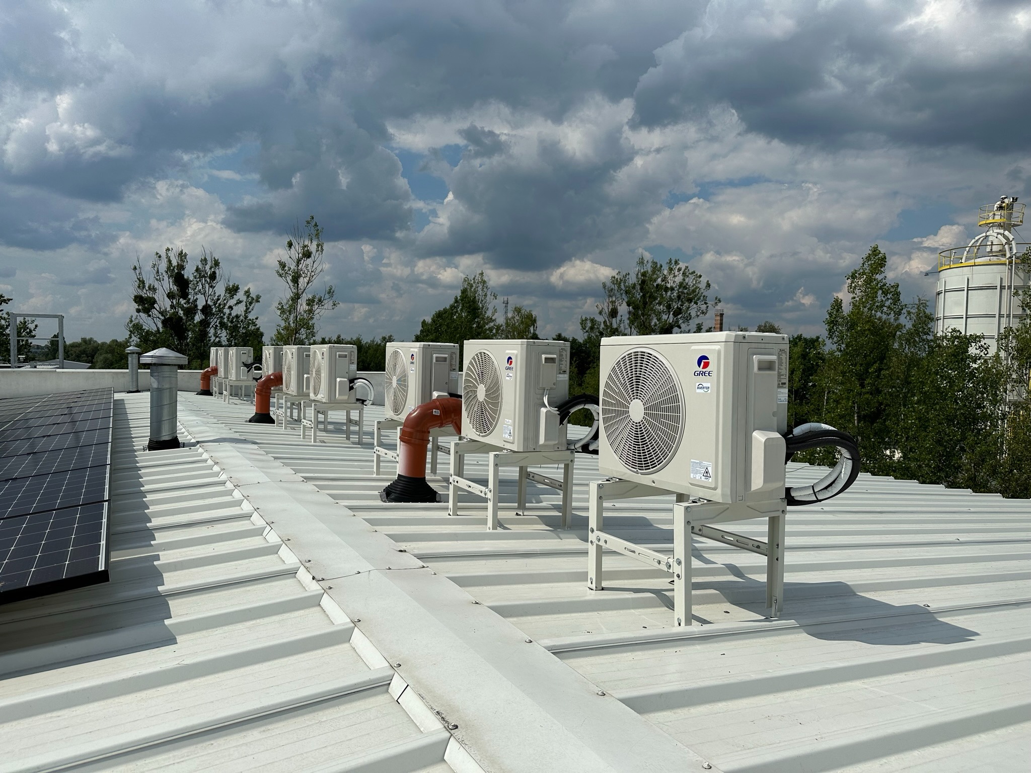 Rząd klimatyzatorów marki Gree na metalowym dachu, z widocznymi panelami fotowoltaicznymi po lewej stronie i elementami instalacji wentylacyjnej.