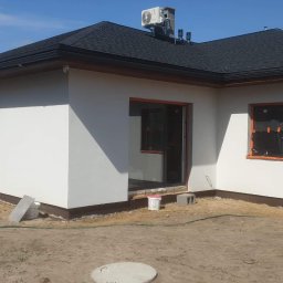 Ariel Kamiński - Malowanie elewacji domu jednorodzinnego z czarnym dachem, widoczny pracownik na drabinie malujący ramę okienną na szaro, okno zabezpieczone folią, obok stoi wiadro z farbą.