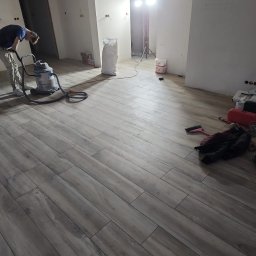 Ariel Kamiński - Wnętrze w trakcie remontu: mężczyzna odkurza nowo ułożoną podłogę z płytek imitujących drewno, widoczne worki z materiałami budowlanymi, oświetlenie robocze i ślady po pracach na ścianach.